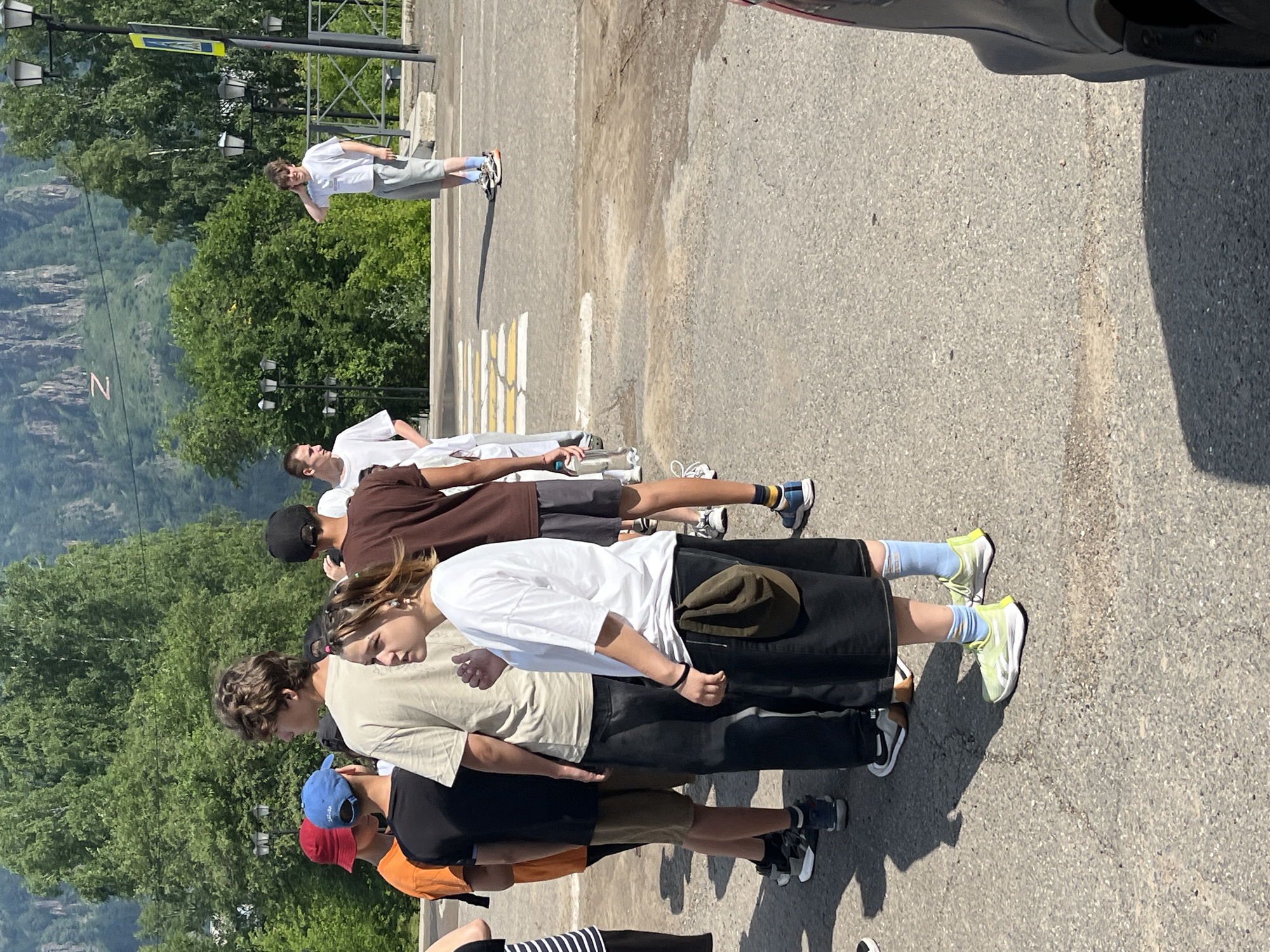 The image depicts a group of young people standing on an asphalt road near a pedestrian crossing, located in the city of Divnogorsk, Russia. The background features lush green trees and rocky mountains, characteristic of the region. Sunlight casts shadows on the ground, indicating it is daytime. The individuals are dressed in casual summer attire, including t-shirts, shorts, and sneakers. One person in the foreground is wearing a black skirt and a white t-shirt. Another individual, a male, is standing to the right, touching his hair. A yellow pedestrian crossing sign is visible on a pole. The overall atmosphere is relaxed, suggesting a casual gathering or a break during an outdoor activity.