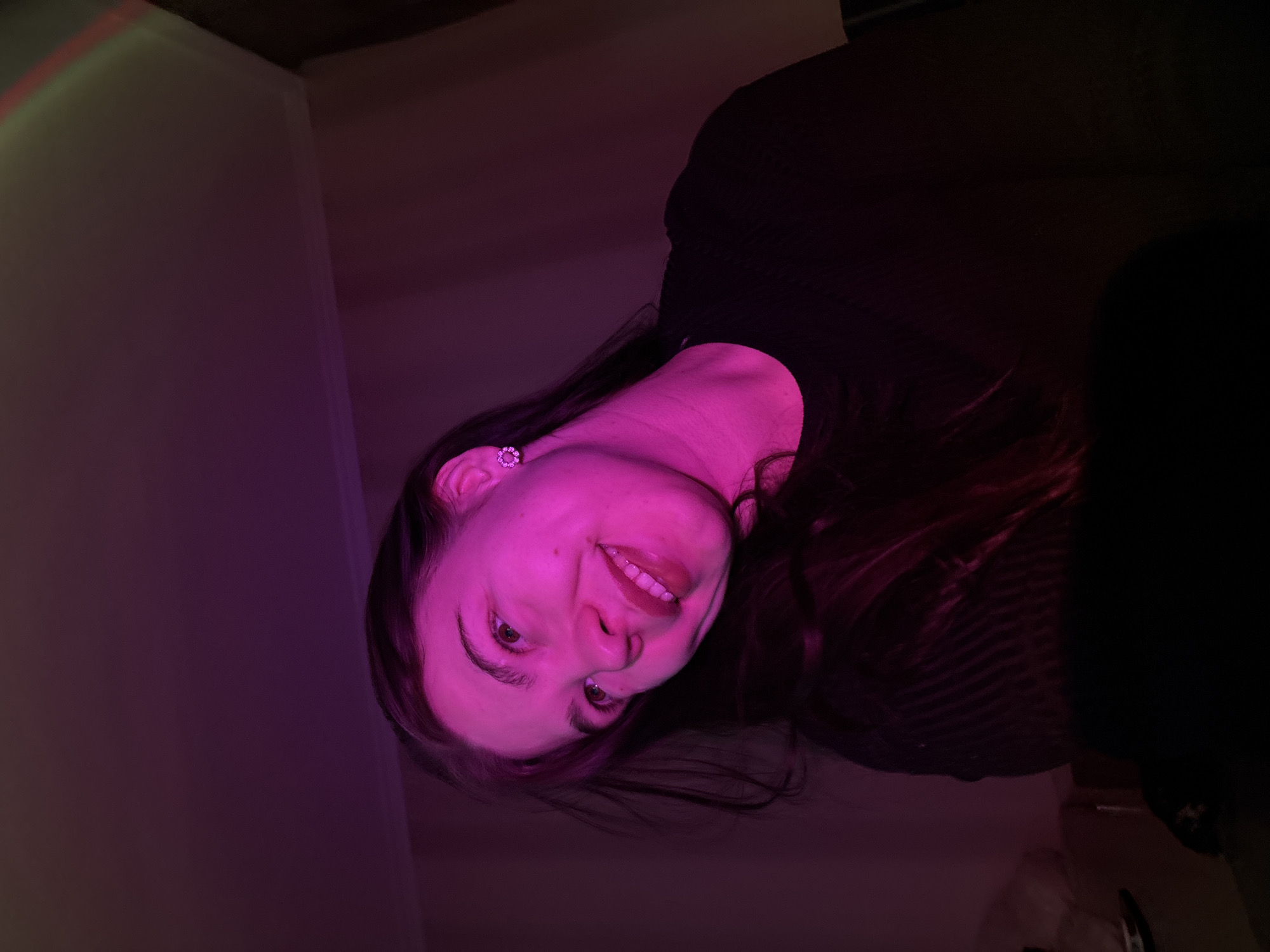 A photograph taken indoors and rotated counter-clockwise shows a young woman with long, dark hair and fair skin under pink lighting. She is wearing a dark-colored top. The background consists of a light-colored wall on the left and dark areas on the right. A floral stud earring is visible on her left ear. The location is unknown, as the image provides no information that confirms it is in Perm, Russia.