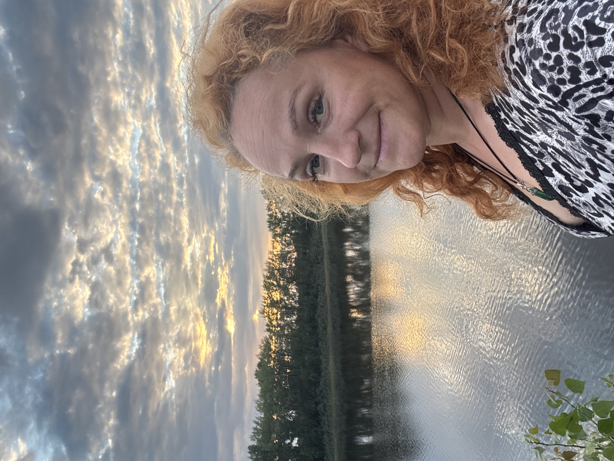 A medium shot, selfie-style photograph is taken outdoors in Chaikovsky, Russia. The subject, a fair-skinned woman with fiery red curly hair, is positioned in the right foreground, looking towards the camera with a slight smile. She is wearing a black and white leopard-print top with black lace trim and a pendant necklace with a green stone. The background features a tranquil body of water reflecting the sky, which is filled with dramatic, cloud-streaked sunset colors of yellow and grey. A dense line of green trees borders the far bank of the water. In the lower left corner, a few green leaves and branches are visible.