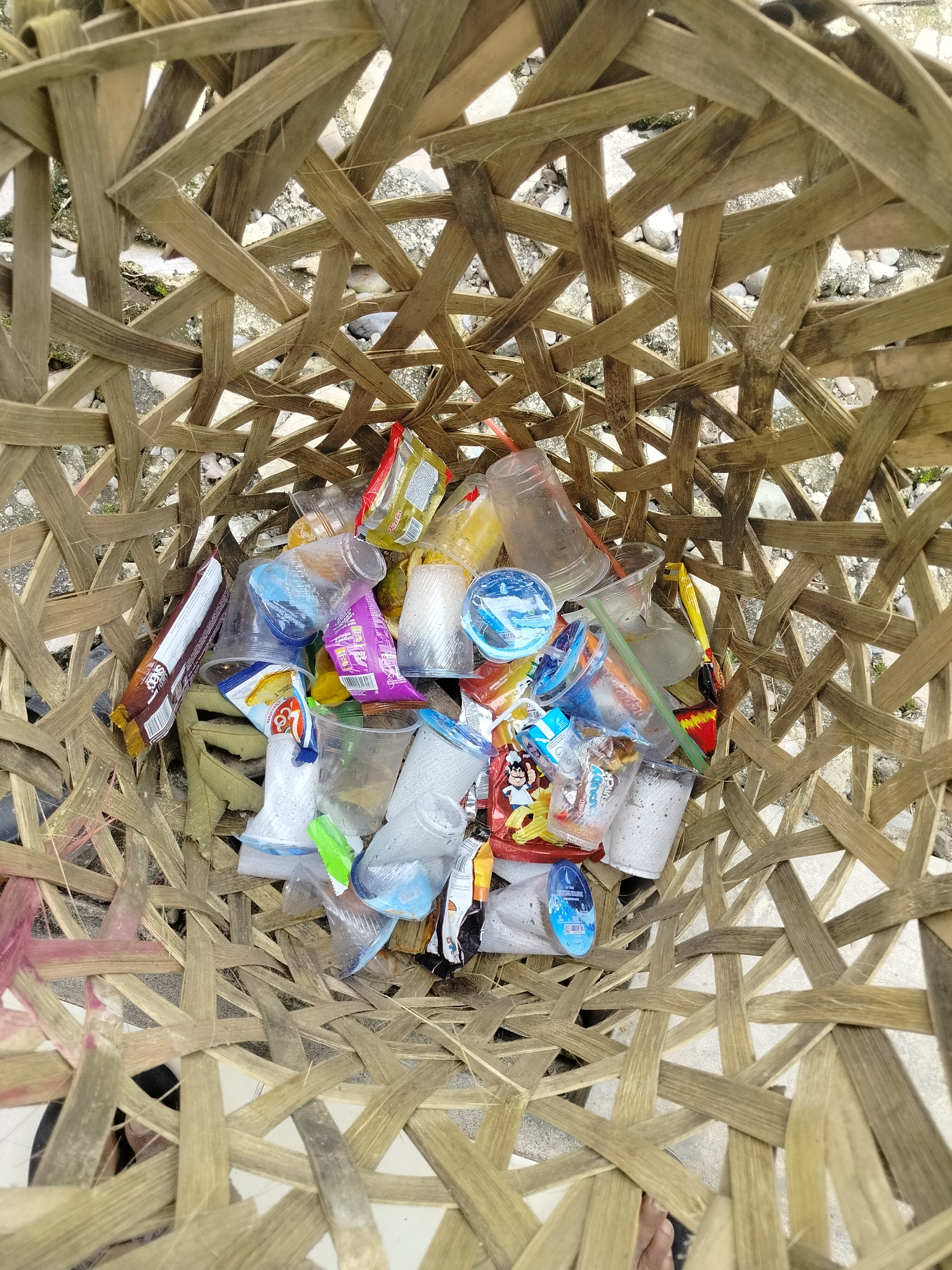 This is a top-down, medium shot photograph of trash within a woven bamboo basket. The basket is approximately 1 meter in diameter and appears to be located outdoors, possibly in an urban area, as indicated by the presence of small rocks and debris on the ground beneath the basket. The contents of the basket consist primarily of single-use plastic items such as cups, wrappers, and small bags, many of which appear to be remnants of snacks and beverages. There are also a few dried leaves mixed in with the debris. The lighting suggests it is daytime, with natural light illuminating the scene. No people are visible in the frame, though a portion of a bare foot can be seen at the bottom right edge of the image, suggesting someone is nearby. The overall impression is one of accumulated waste in a rustic receptacle. The location is identified as Purwodadi, Indonesia, a city known for its agricultural sector and natural attractions.
