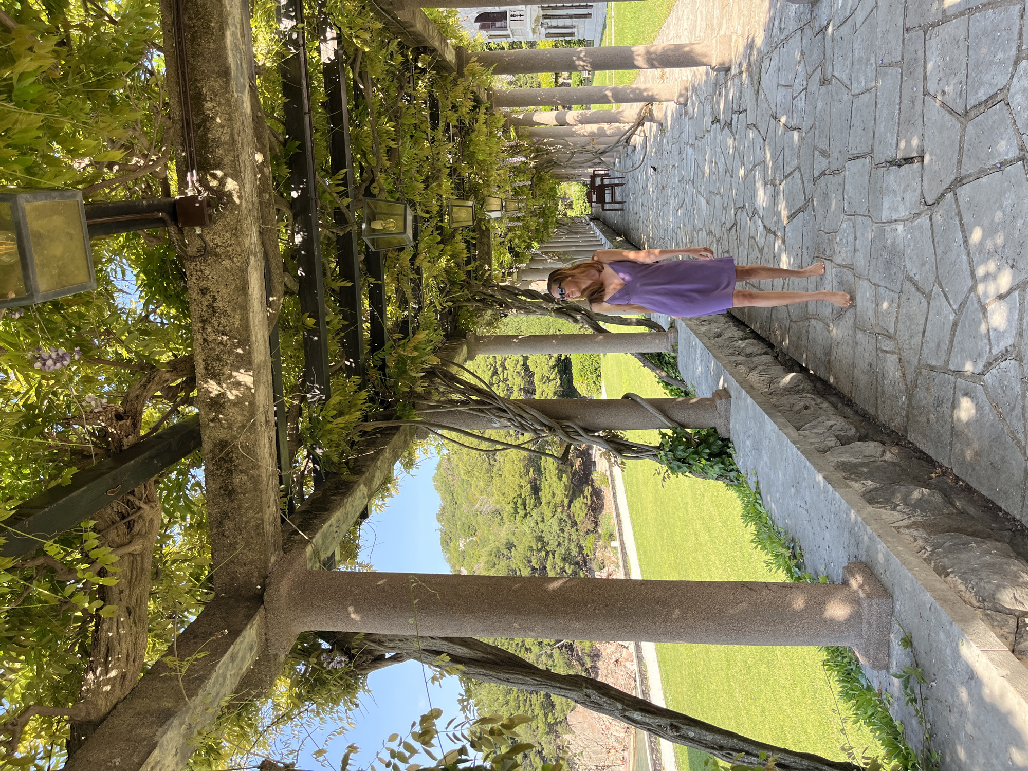 This image, originally taken in portrait orientation and rotated 90 degrees clockwise, depicts a woman strolling barefoot along a stone pergola at Sveti Stefan, Montenegro. The pergola is constructed with sturdy grey stone pillars and beams, enveloped by abundant green wisteria vines, some bearing subtle purple blossoms. Ornate, square-shaped glass lanterns hang from the structure, with electrical wiring and a small junction box visible on one of the pillars.
The pathway beneath the pergola is paved with irregular grey flagstones, highlighted by bright sunlight and scattered shadows. A woman with light brown hair, wearing sunglasses and a knee-length purple dress, stands relaxed on the path, looking towards the viewer. Her bare feet emphasize the warm, leisurely ambiance. Further down the path, a wooden table and chairs are discernible.
Beyond the pergola, a neatly manicured green lawn leads to a dense, forested hillside under a clear blue sky. The strong sunlight and distinct shadows suggest a bright, warm day, likely late morning or early afternoon. The scene evokes a tranquil and picturesque setting, characteristic of a historical or resort location. No visible text is present.