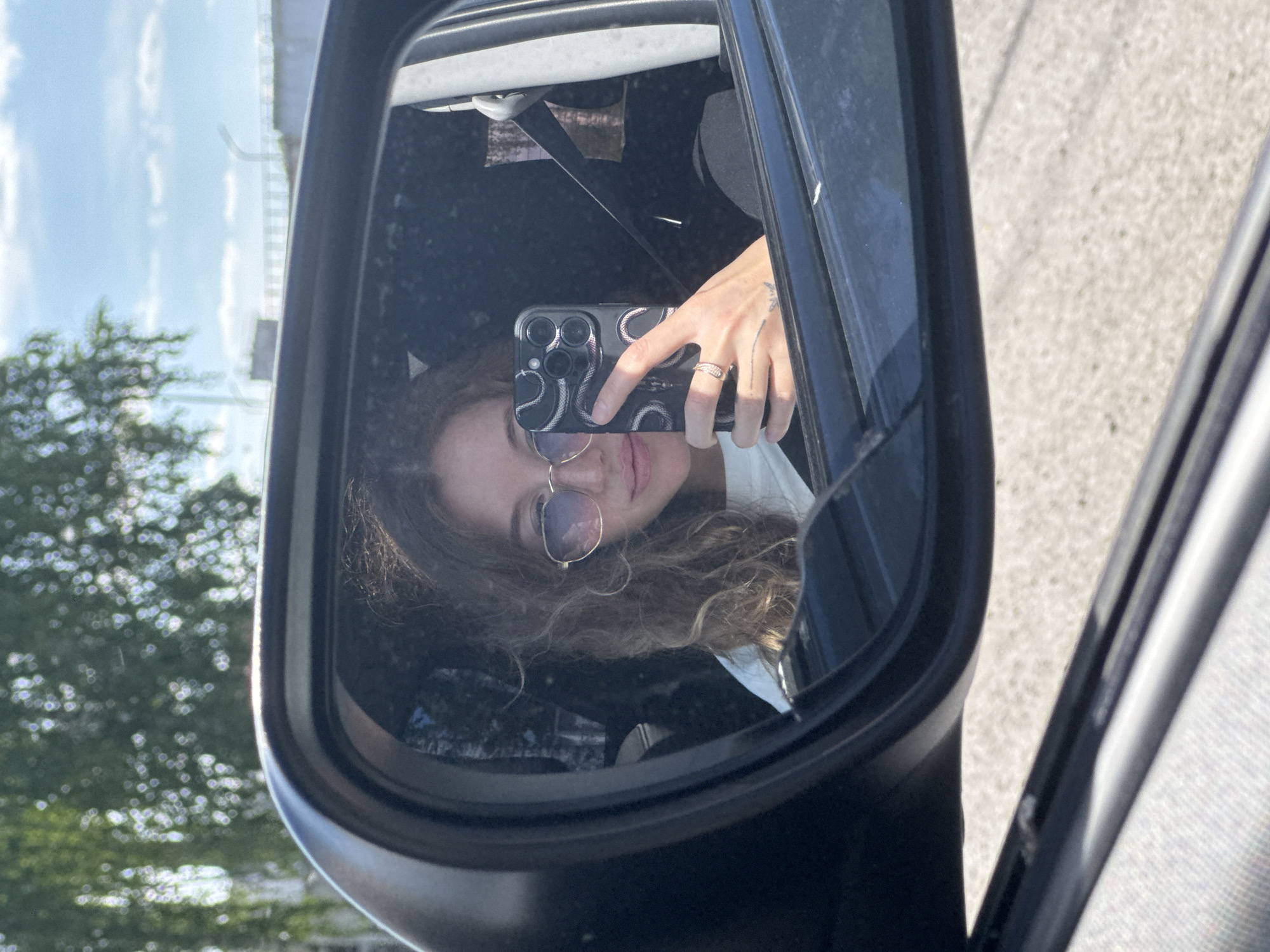 The image displays a young woman taking a selfie reflected in the driver's side exterior mirror of a vehicle, in the city of Chaikovsky, Russia, under bright daylight conditions.
The woman has long, wavy brown hair and is wearing round, light-tinted sunglasses and a white top. She holds a dark-colored smartphone with her right hand, which features a distinctive black case with a white and pink serpentine pattern and a multi-lens camera array. A gold-toned ring is visible on her right ring finger, and a small, dark tattoo is present on the back of her right hand near the wrist. Her lips are slightly parted.
The car's black exterior mirror casing frames her reflection. Within the mirror, behind her, the reflection shows a clear blue sky with scattered white clouds, and out-of-focus green foliage from trees, indicating an outdoor setting. Faintly visible beyond the trees are light-colored concrete structures, possibly a bridge or elevated roadway infrastructure.
Elements of the car's interior are also visible: a dark seatbelt strap runs across the upper right portion of the reflected image, and a glimpse of a light grey textured fabric, likely a door panel, is seen in the bottom left corner outside the mirror's frame. The ground directly below the mirror and the car's exterior frame appears to be a light grey paved surface, consistent with asphalt or concrete, illuminated by direct sunlight.