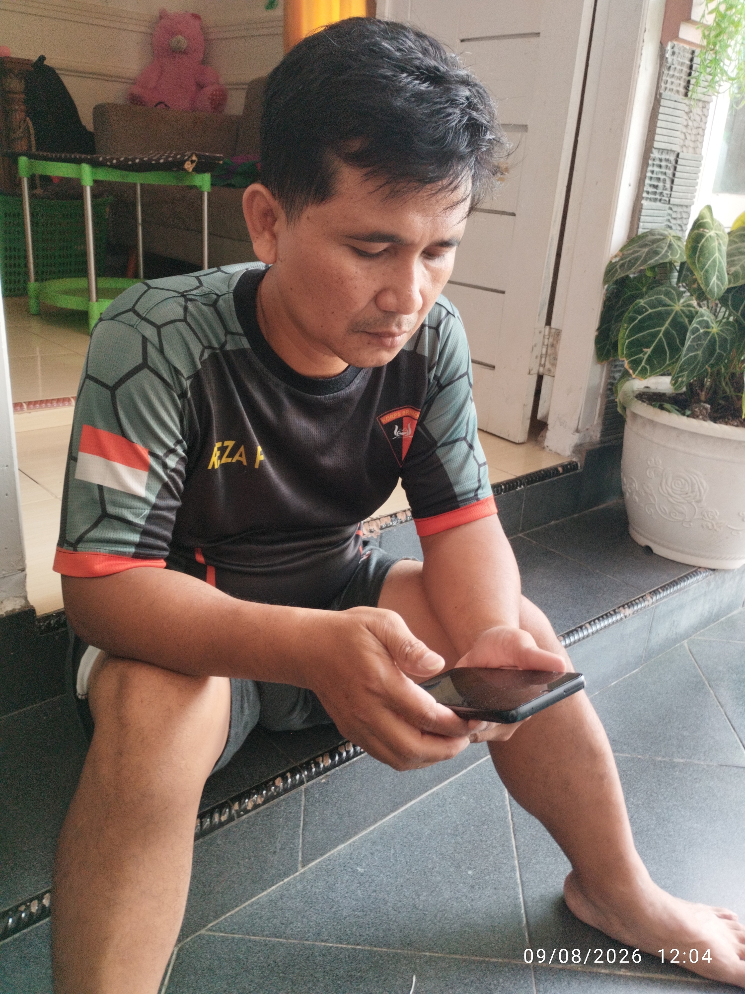 The image shows a man sitting on dark grey tiled steps outdoors, looking down at his smartphone. He is wearing a grey and black t-shirt with a honeycomb pattern on the sleeves, an Indonesian flag on his left sleeve, and the text "REZA P" on the front. He is also wearing dark grey shorts. His right leg is visible, with light hair. To the right of the man, a potted plant with large, dark green leaves is visible. The background shows a doorway with a white-painted frame and a hint of a window. In the upper left background, a pink teddy bear is sitting on a couch. The scene appears to be a residential area in Bandar Lampung, Indonesia. A timestamp in the lower right corner indicates the date 09/08/2026 and time 12:04.