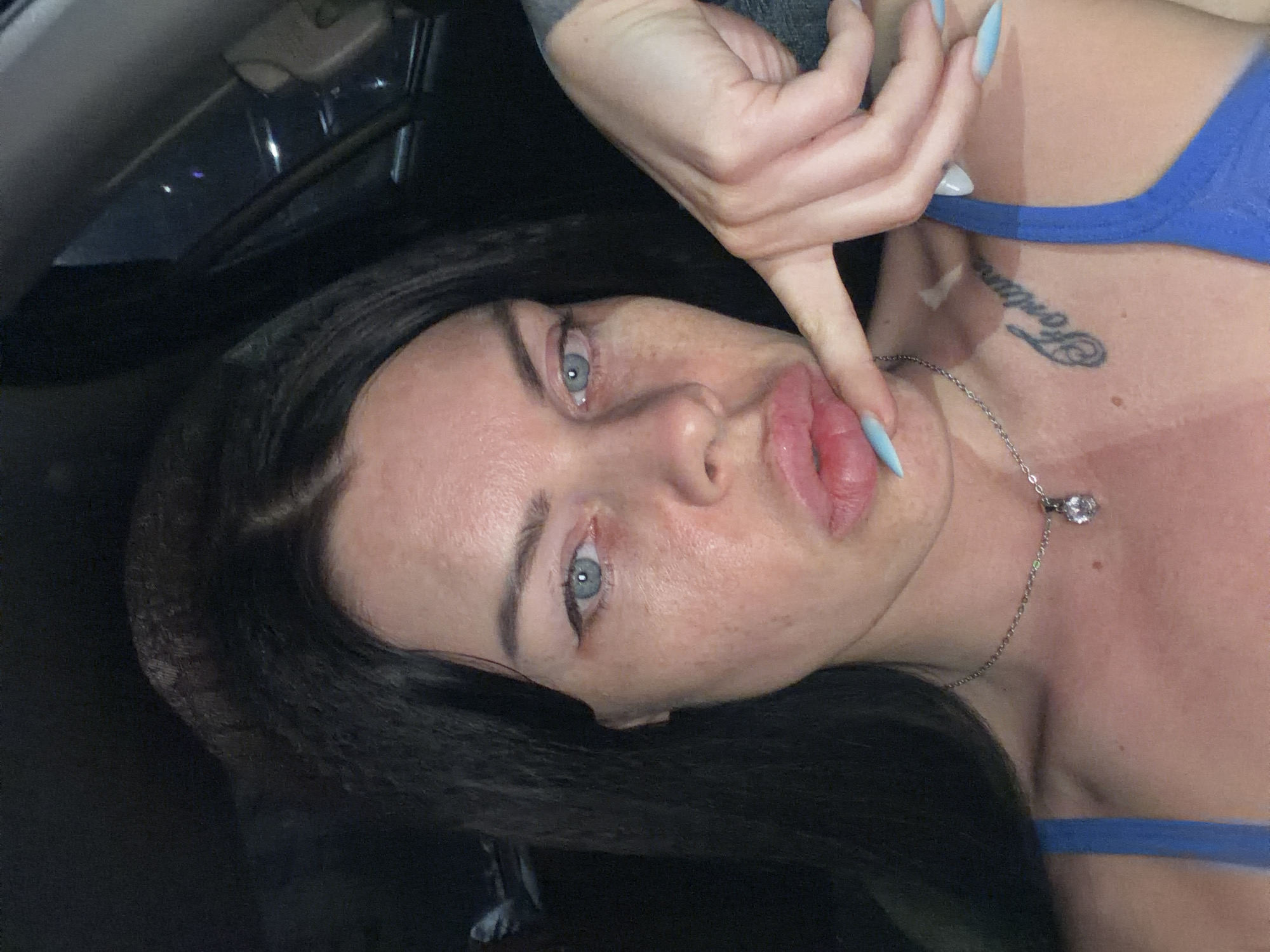 The image shows a close-up, vertically oriented portrait of a woman with dark hair and light blue eyes. She is wearing a blue top and a silver necklace with a clear pendant. A tattoo is visible on her neck, and she has long, light blue-tipped fingernails. Her right hand is positioned near her face, with her index finger touching her lips. The lighting suggests an indoor setting, possibly a car, with some darker areas indicating a shadowed environment. The woman appears to have light makeup on, and her skin shows some texture. There are no clear indicators of the time of day or weather. The context of Ufa, Russia is provided, but not visually evident in the image itself.