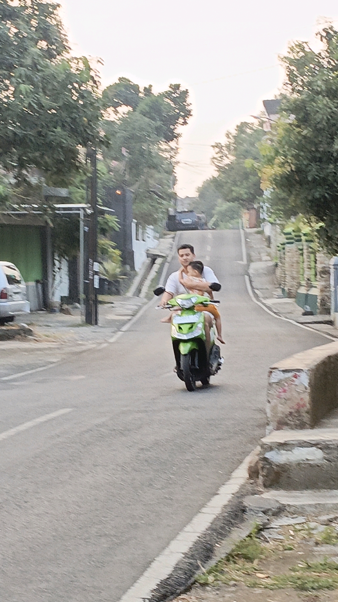 In Salatiga, Indonesia, a man is riding a green scooter up a paved street. He is wearing a white t-shirt and is holding a young child in his arms who is wearing orange shorts. The child is facing forward, with their arms around the man's neck. The scooter has a white front panel and a black tire visible. The street is lined with trees and buildings on either side. There are power lines visible above the trees. The sky is overcast.