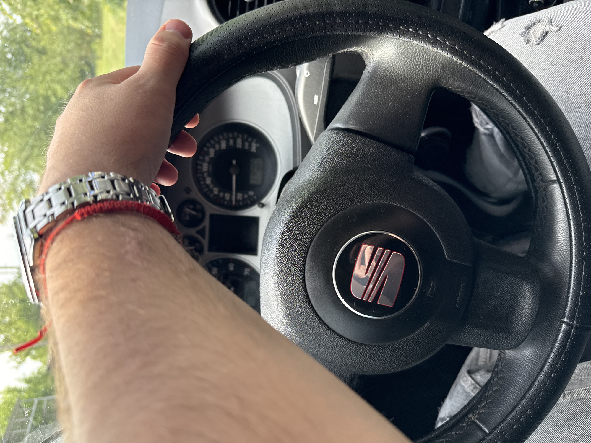 A first-person perspective image captures a person's right hand and forearm gripping a black leather-wrapped steering wheel in a car during the daytime. The hand, with visible arm hair, wears a metallic link wrist watch and a red string bracelet. The steering wheel, likely showing signs of use with slight wear on its textured surface and outer rim, features a red and metallic SEAT logo centrally on its hub. The word "AIRBAG" is visible on the right spoke of the steering wheel.
Behind the steering wheel, the car's instrument cluster is partially visible, showing a speedometer with numerical markings (e.g., 20, 40, 60, 70, 80) and a needle pointing towards a low speed or zero, suggesting the car is stationary or moving slowly. Through the windshield, outside the car, bright green foliage from trees and a bright sky indicate a clear, sunny day. The person's leg, wearing distressed light-colored denim jeans, is partially visible in the lower right corner of the frame. The overall scene depicts someone inside a vehicle, possibly in Donets, Ukraine, preparing to drive or paused during a journey.