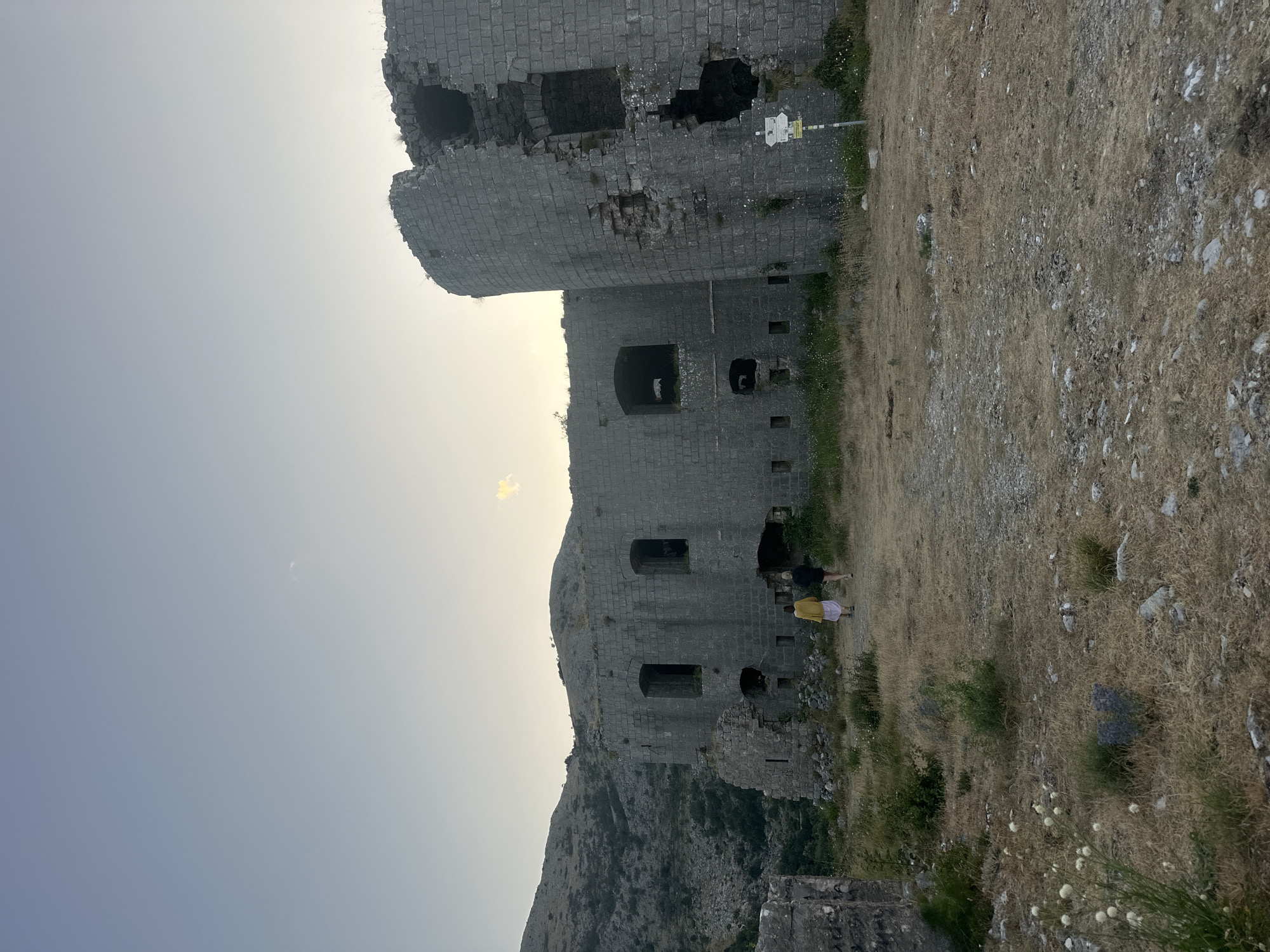In Brajici, Montenegro, a low-angle, full-shot photograph captures a dilapidated stone fortress under a clear, pale sky. The fortress, constructed from large, grey stone blocks, exhibits significant structural damage, with gaping holes and crumbling sections, particularly on its rounded turret. Two individuals, one wearing a yellow jacket and the other a dark top and shorts, are walking away from the camera towards the fortress. They are positioned in the mid-ground on a dirt path. The foreground consists of dry, golden grass and scattered stones, with a few small white flowers visible. The background features a rugged, mountainous landscape with sparse vegetation. A yellow sign with directional arrows is affixed to a pole near the fortress wall. The lighting suggests late afternoon or early evening.