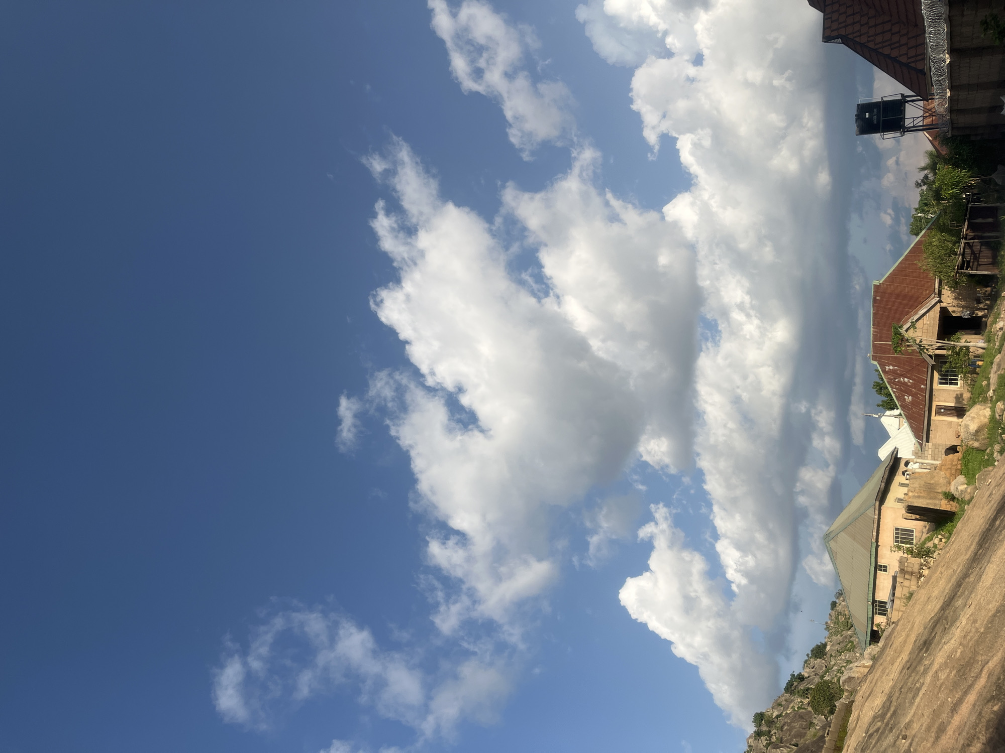 The image portrays a residential scene in Barakin Akawo, Nigeria, featuring natural rock formations and human structures under a daytime sky. The upper two-thirds of the frame shows a clear blue sky interspersed with numerous white cumulus clouds, casting shadows and highlights indicative of direct sunlight.
The left foreground and midground are dominated by a large, smooth, reddish-brown natural rock slope. Above this, a rocky hill with grey boulders and sparse green shrubs and trees rises.
Multiple single-story houses are visible. In the left-midground, a building with light-colored walls and a dark green gabled roof is positioned on the rock slope. This house has several rectangular windows, some fitted with security bars. A single individual, dressed in light-colored clothing, is standing near the front of this house, oriented towards the right side of the frame.
To the right, another single-story house features light-colored walls and a rust-colored corrugated metal roof. A black, cylindrical water tank on a metal stand is visible to the right of this house. Further right, the top section of a third house with a dark reddish-brown corrugated metal roof is partially visible, behind a wall topped with barbed wire fencing. Green foliage is scattered around the properties and on the rocky terrain.