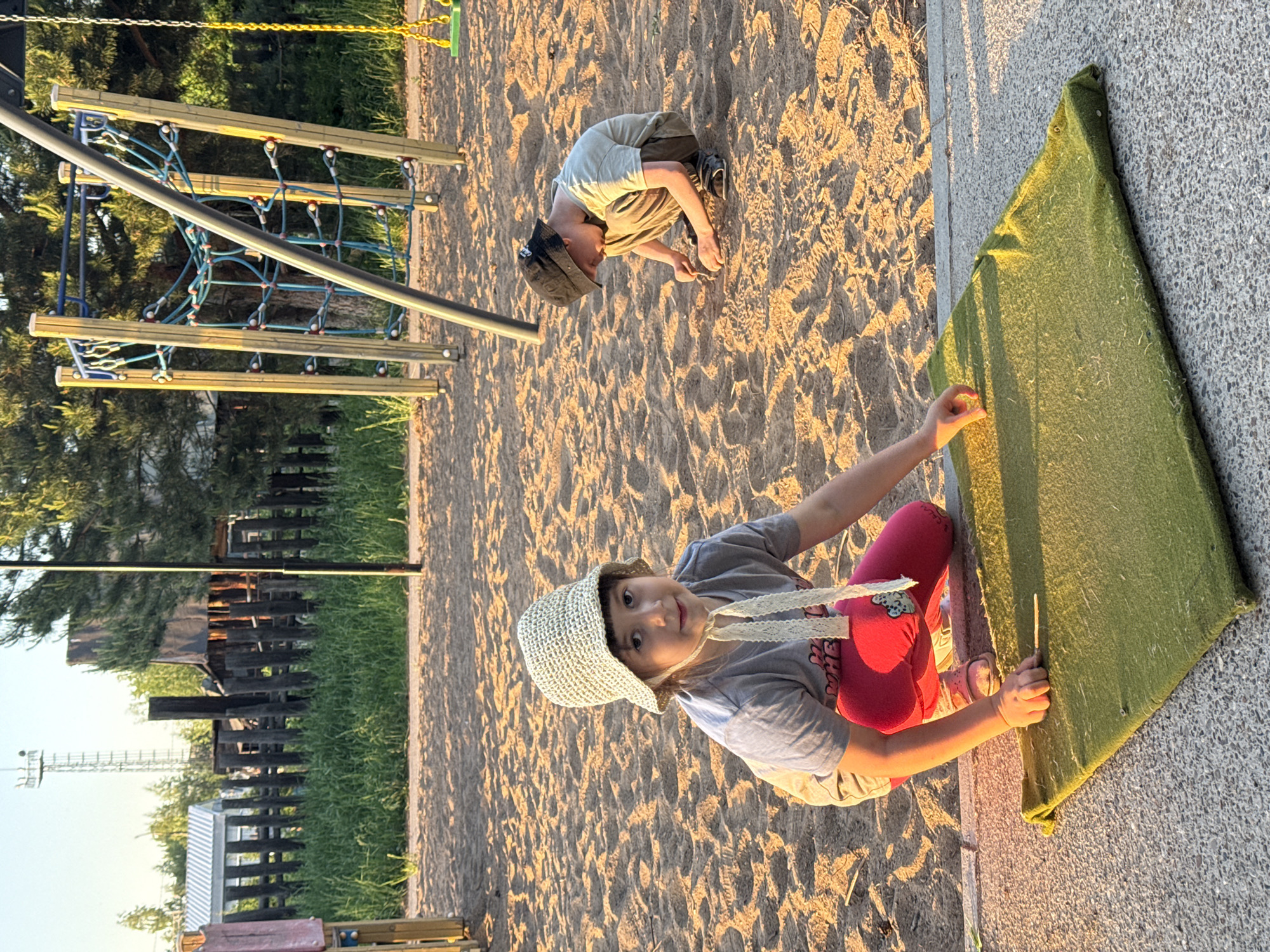 The image shows two children on a playground, likely located in Chaikovsky, Russia given the prompt instructions. A girl in a beige sunhat, gray T-shirt, and pink leggings is kneeling on a sandy surface near a gray concrete wall. She is touching a green, possibly padded, panel attached to the wall. A boy in a dark cap and light-colored shirt and pants is kneeling in the sand in the background, seemingly playing. In the background there is also a metal playground structure with wooden beams and blue ropes. The playground appears to be near a grassy area with a fence and some buildings. The lighting suggests it is late afternoon or early evening.