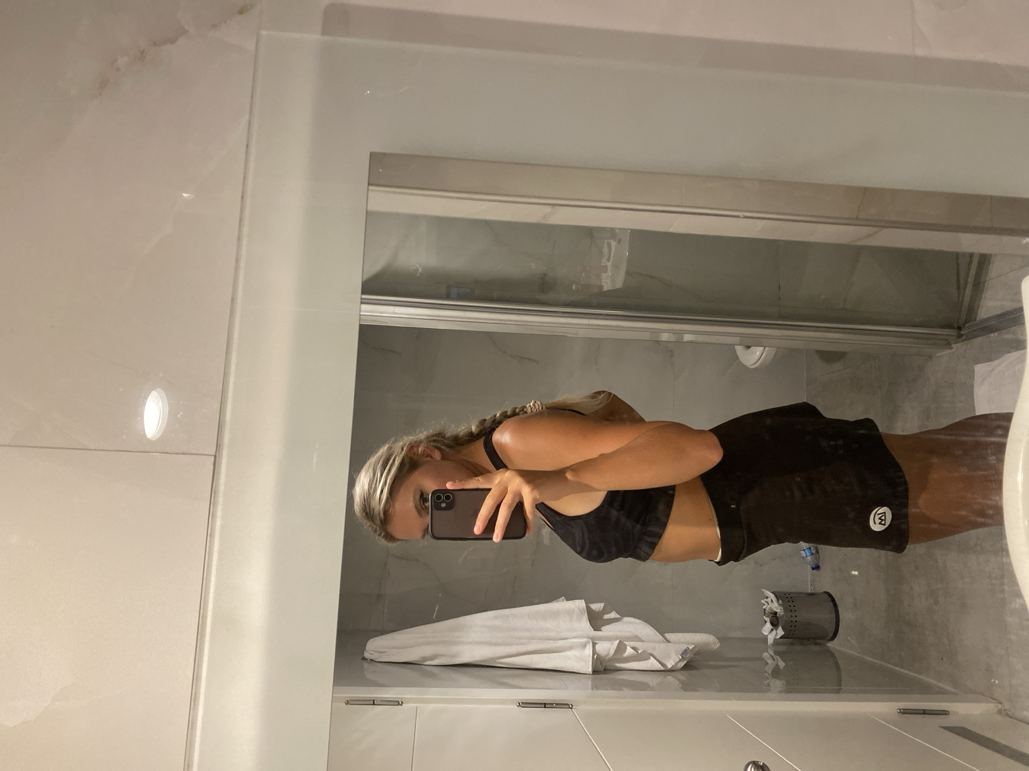 A woman with blonde hair in a braid is taking a mirror selfie in a bathroom. She is wearing a black sports bra and black shorts. The bathroom appears to be modern, with marble-like tiles on the walls and a glass shower door. A white towel is hanging on a rack to the left of the woman. A silver trash can with some items in it is visible in the reflection of the mirror. The scene takes place in a bathroom located in the city of Aksu, Turkey.