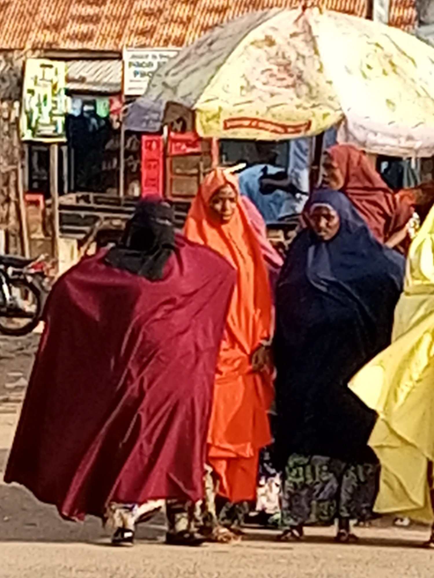 The image shows a group of people dressed in traditional attire, primarily women in colorful hijabs and abayas. They are standing in what appears to be an outdoor marketplace or street scene. The background includes stalls with various items displayed, suggesting a commercial area. A motorcycle is partially visible on the left side of the frame, indicating a mode of transportation commonly used in the area. The lighting suggests it is daytime, likely sunny, given the bright colors and shadows. The overall atmosphere seems casual and everyday, with no particular event clearly depicted. Some of the visible text on signs in the background is blurry and difficult to decipher, but one sign appears to have large block letters, possibly related to shop names or advertisements. The clothing suggests a cultural context where modesty is observed. The people's faces are not clearly visible, obscuring any specific emotions they might be displaying. The scene is likely set in Tempure, Nigeria, as suggested by the provided location context, which implies a West African setting.