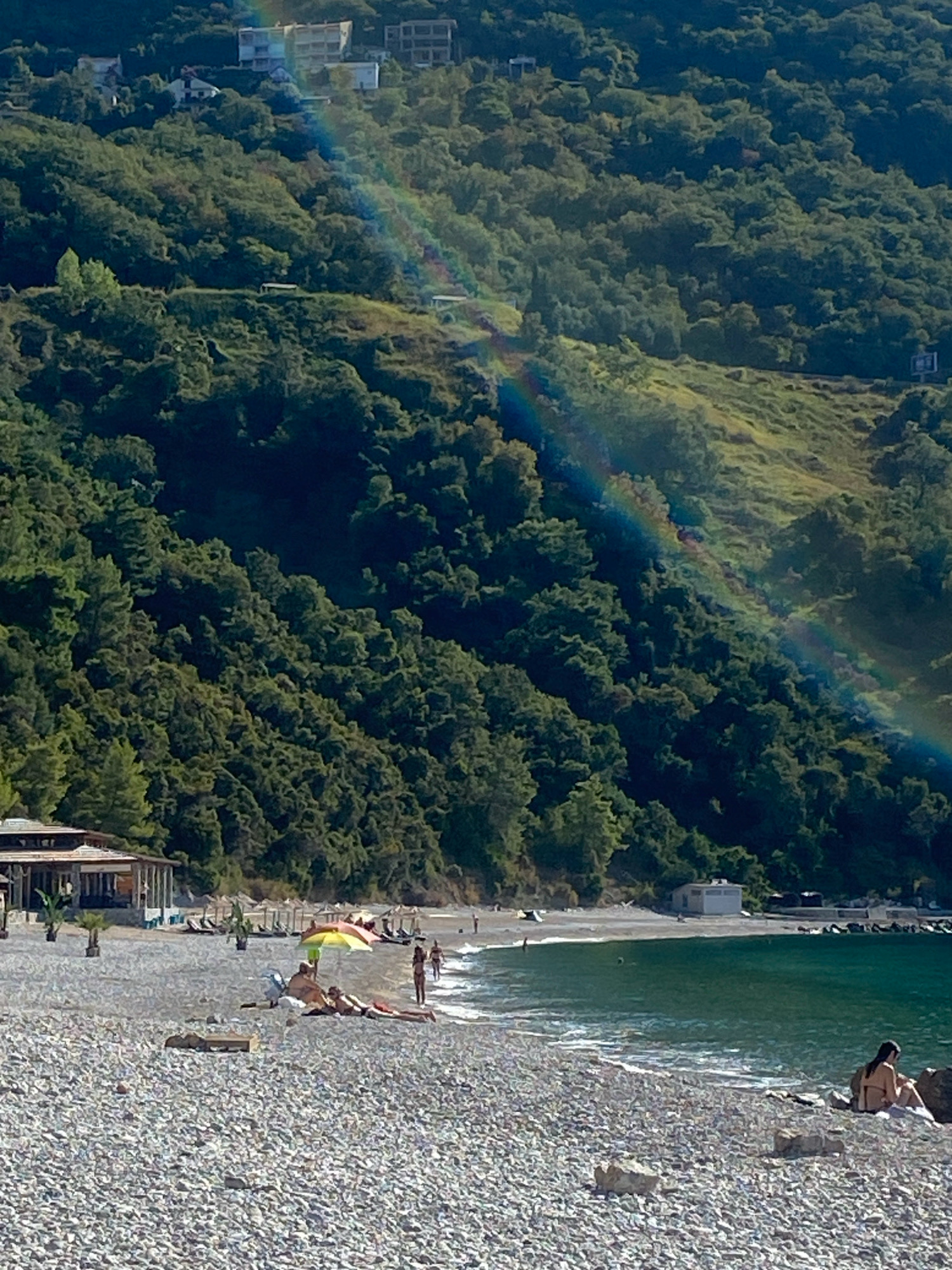 The image depicts a pebble beach in Prijevor, Montenegro, with a lush green hillside in the background. A faint rainbow arcs across the hillside, intersecting with some buildings visible at the top. On the beach, several people are seen relaxing. Some are lying on towels, while others are sitting or walking. One person is sitting on rocks near the water's edge. Umbrellas provide shade on the beach, and a small building, possibly a beach bar or restaurant, is visible on the left side of the image. The sea is a deep, vibrant blue-green, with gentle waves lapping at the shore. The overall scene conveys a sense of leisure and natural beauty.