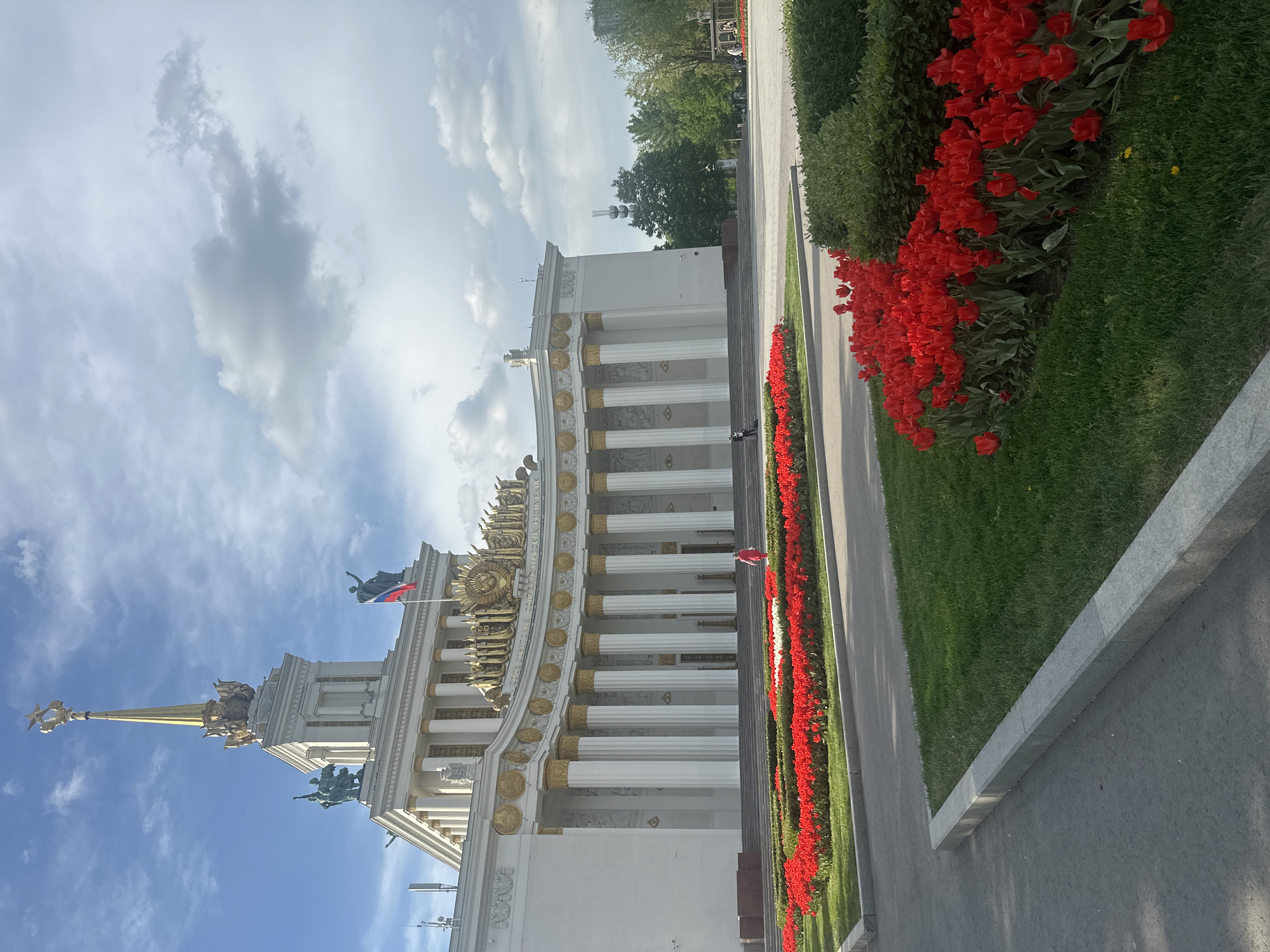 The image shows the main pavilion of the Exhibition of Achievements of National Economy (VDNKh) in Moscow, Russia. The building is white with gold detailing and classical architectural elements like columns. A Russian flag flies from the top. Along the side of the building is a row of red tulips. The sky is blue with clouds.