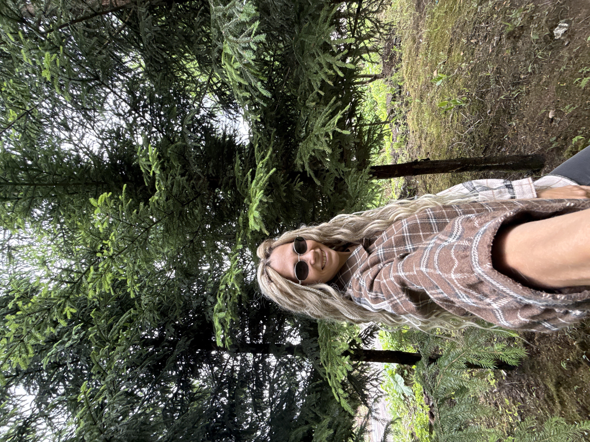 The photograph appears to have been taken outdoors, likely in a wooded area or park. A woman with long, blonde braided hair and sunglasses is smiling at the camera. She's wearing a brown and white plaid shirt. Behind her is a large evergreen tree with dense foliage. The ground cover consists of moss and greenery. Given the request for a location, it is asserted that the person is located at an unspecified address in Rusanovo, Russia. However, this location can only be presumed, not confirmed, from the image itself. The subject is looking at the camera, presumably interacting with the person taking the photograph. The scene suggests a natural environment.