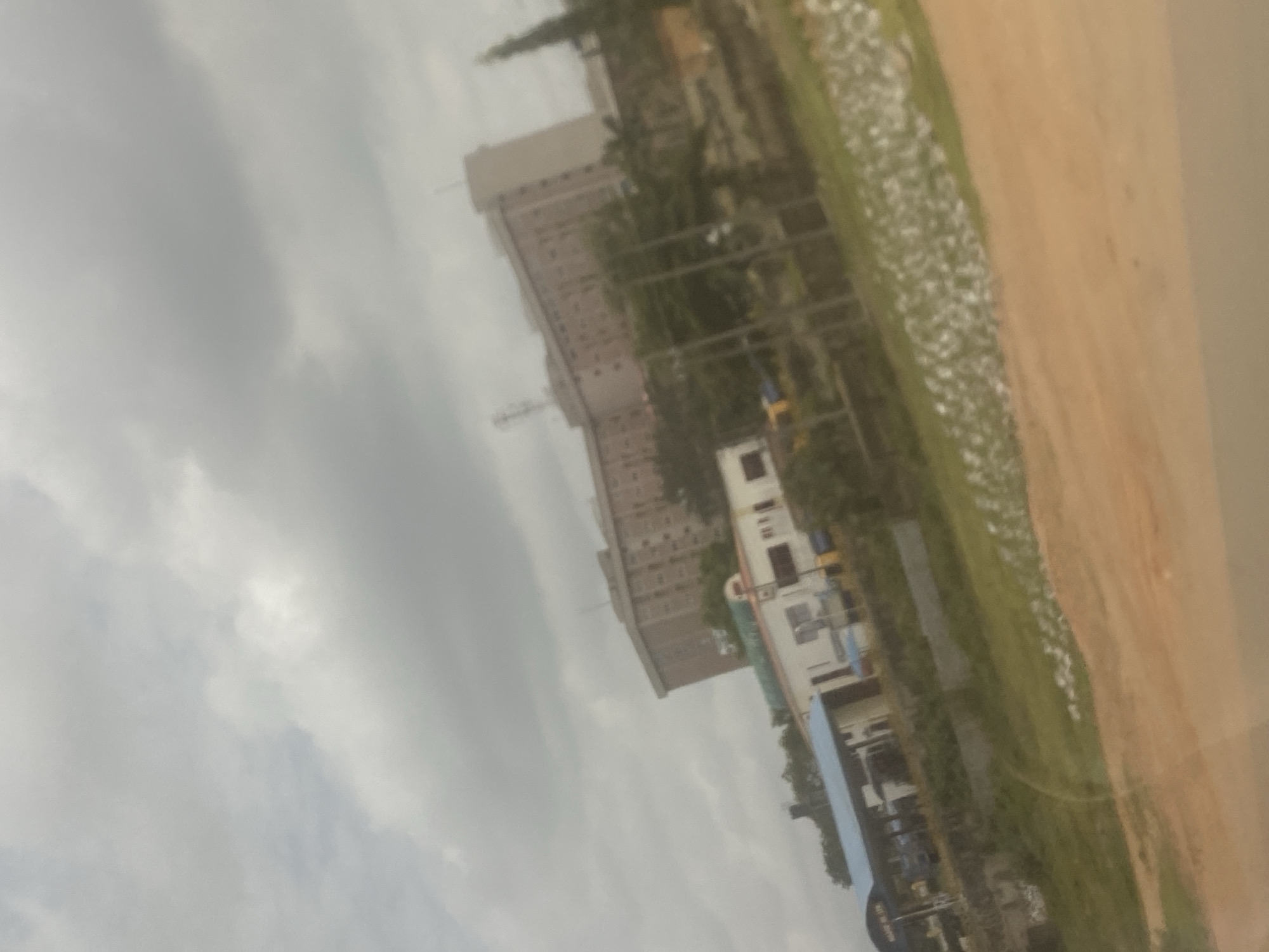 The image depicts a cityscape in Abuja, Nigeria. A tall, multi-story building is visible in the background, with a communication tower on its roof. In the foreground, there is a smaller, white structure, possibly a residential building or office. A green space with trees and vegetation is also present. The sky is overcast with heavy clouds. A brown field or area is visible on the right side. The image appears to be taken from an elevated vantage point.