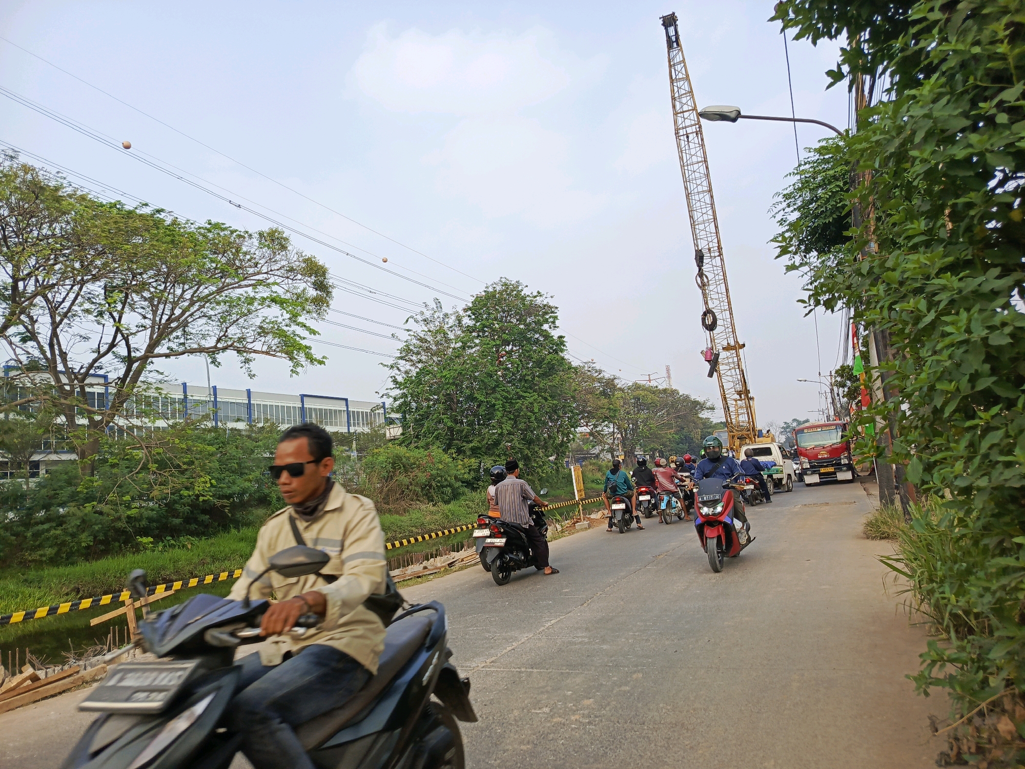 The image depicts a street scene in Tangerang, Indonesia, under an overcast sky. In the foreground, a man wearing sunglasses and a light-colored long-sleeved shirt rides a dark-colored motorcycle. He is looking towards the left side of the frame. Behind him, a line of motorcycles and some cars are proceeding down the road. Several riders are visible on the motorcycles. In the background, a tall yellow construction crane dominates the skyline, indicating ongoing construction or infrastructure work. Trees and greenery line the sides of the road, and a building with a modern facade is visible on the left. The road surface appears to be asphalt, with some markings. A red truck is also present in the traffic. The overall atmosphere suggests a typical day in a developing urban area.