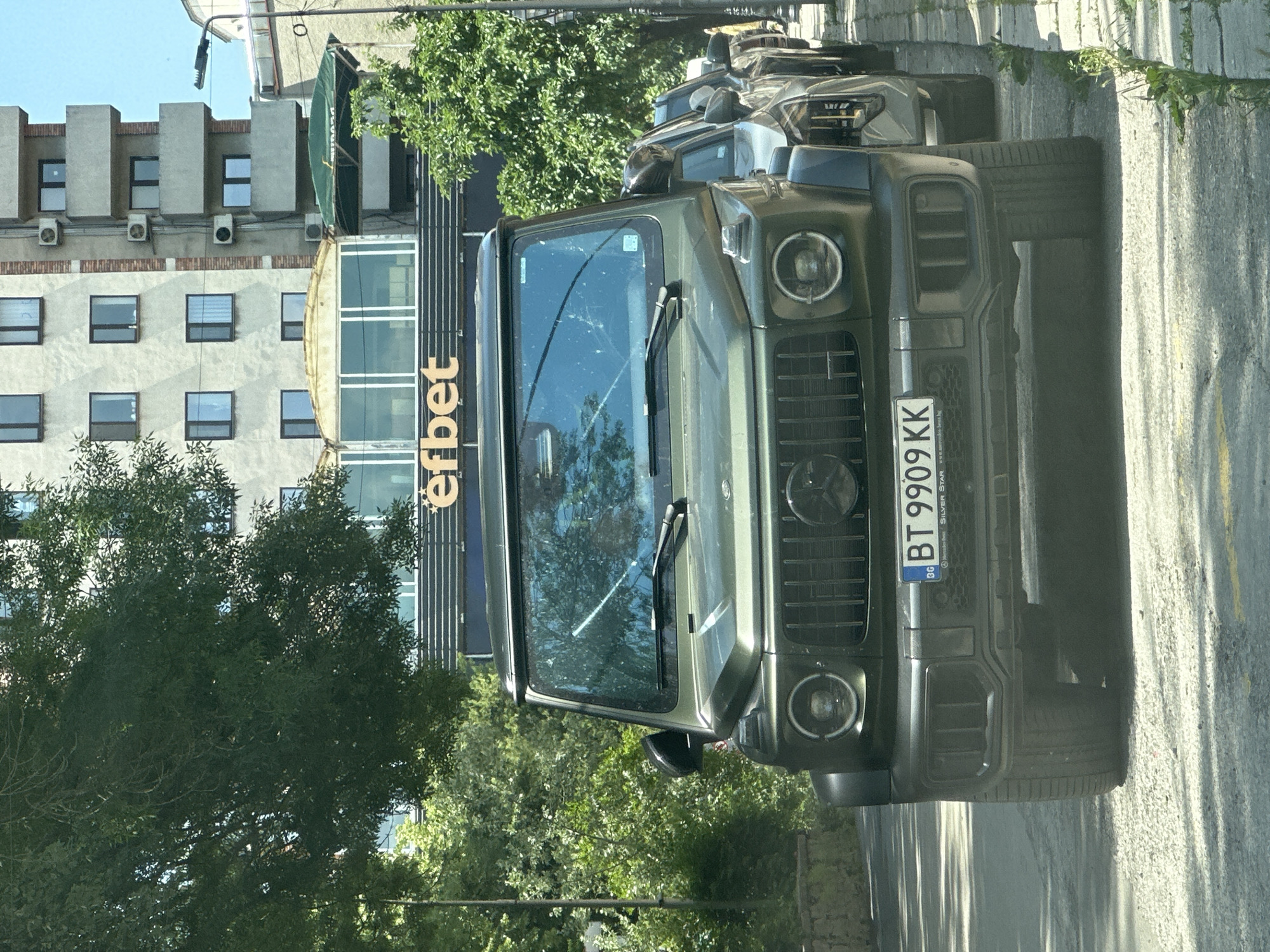 The image depicts a green Mercedes-Benz G-Class SUV parked on a street in Veliko Tarnovo, Bulgaria. The vehicle's license plate number is BT 9909 KK. In the background is a building with the "efbet" logo. Trees are present on either side of the scene. There is a building behind the Efbet building. The image is taken from a slightly low angle, showing the front of the car. Another vehicle is parked behind the SUV. The environment appears to be a typical urban setting with buildings and greenery.