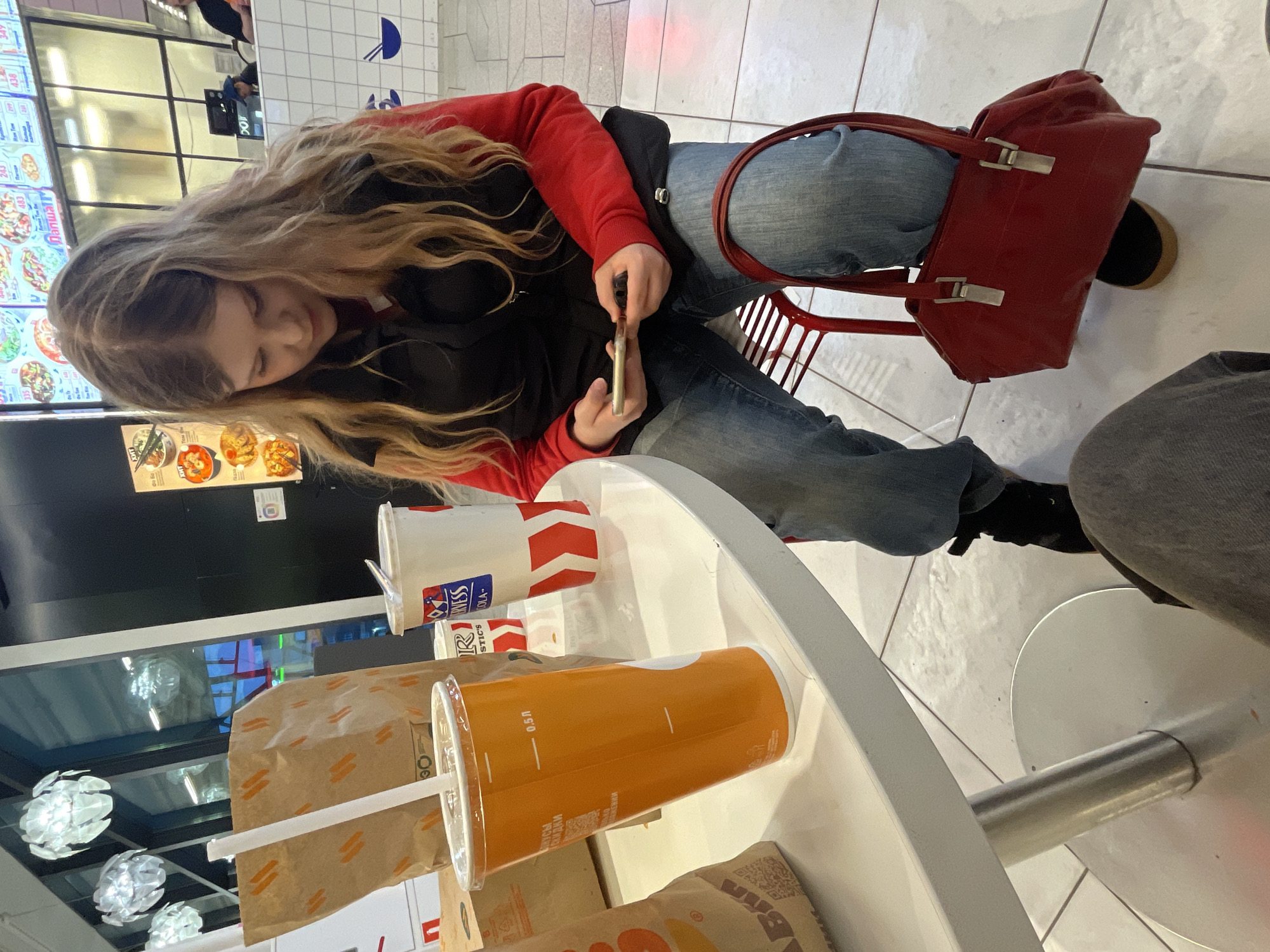 A young woman with long blond hair sits at a round white table, holding a gold-colored object. She wears a red sweater over a black vest, and jeans. A red purse hangs off the chair behind her. On the table are a large orange cup with a white straw, a white and red cup, and several brown paper bags, possibly containing food. Another person sits to the right, only their grey pant leg visible. The setting appears to be a fast-food restaurant or food court. A menu board is visible in the background, as are other tables and chairs. Based on the Cyrillic writing on the cups, the restaurant is located in the city of Saint Petersburg, Russia.