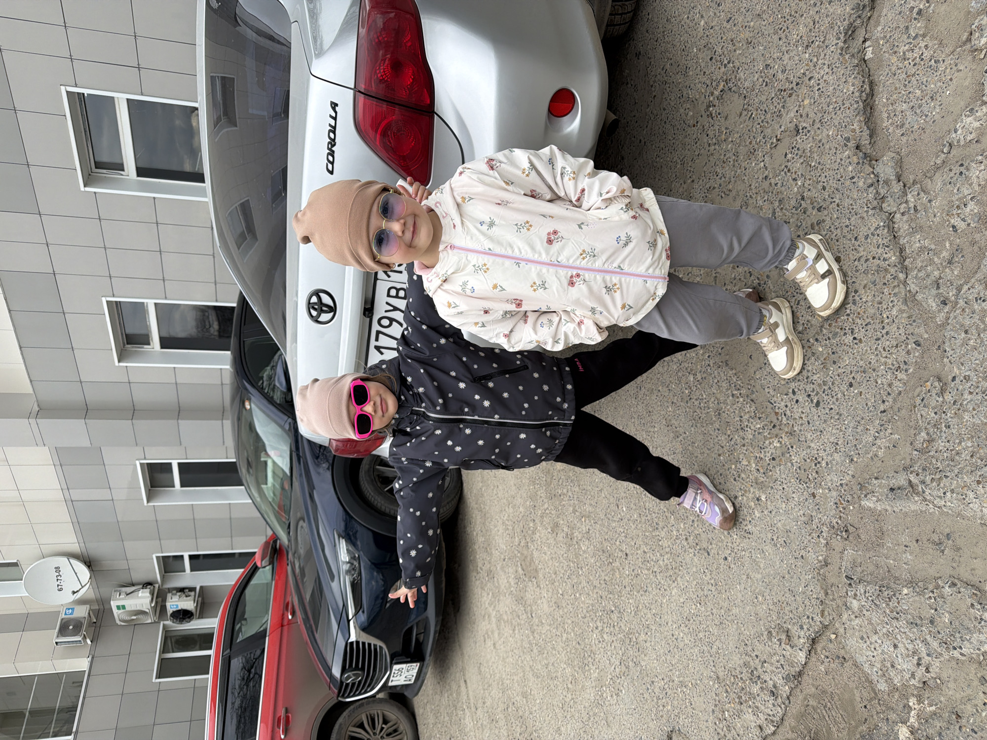 Two young girls, dressed in cool-weather attire, stand side-by-side on an asphalt surface in what appears to be a parking area in Chaikovsky, Russia. The image is oriented sideways, but depicts the girls standing upright.
The girl on the left wears a black jacket adorned with white star-like patterns, black pants, and light purple sneakers with white soles. She has a light brown beanie on her head and is sporting bright pink, square-framed sunglasses. Her left arm is extended outwards, and she looks towards the camera with a slight smile visible behind her shades.
The girl on the right is dressed in a light cream-colored jacket with a delicate floral pattern and grey pants. She wears light brown and white sneakers, a light brown beanie similar to the other girl's, and round, light purple-tinted sunglasses. She also looks towards the camera, seemingly smiling.
Behind the girls, several cars are parked. Directly behind the girl on the right is the rear of a silver Toyota Corolla. The car's tail lights are visible, along with the "COROLLA" emblem and the Toyota logo. Part of its license plate, "179YB 1..", is discernible. To the left, behind the other girl, is the front section of a dark blue SUV, with a partial view of its license plate: "T 556 AO 152". A red car is also partially visible further to the left.
In the background, a modern building with grey paneled walls and multiple windows is visible. Mounted on the building are a white satellite dish with the text "G7-73-08" and several outdoor air conditioning units. The ground is a mix of asphalt and concrete, showing various cracks and patches. The overall impression is that of a casual outdoor moment captured on a possibly overcast day, given the soft lighting.