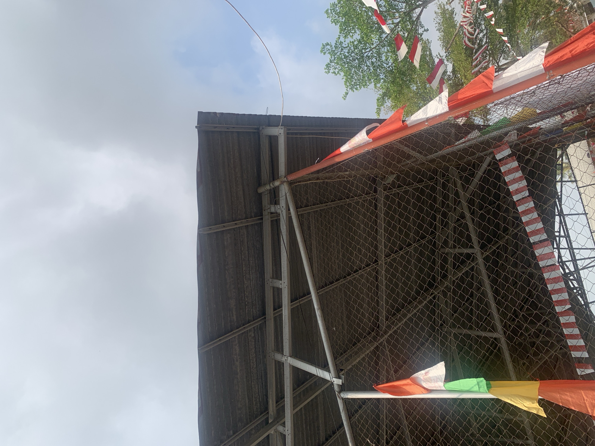 The image depicts an outdoor scene with a clear sky and some clouds. In the foreground, a chain-link fence encloses an area beneath a corrugated metal roof supported by a metal frame structure. Several flags are visible, including a tall pole with multiple flags of various colors (orange, red, white, green, yellow) and a string of red and white pennant flags draped along the fence and the roof structure. To the right, trees with green foliage are visible against the sky. A thick cable runs horizontally across the upper right portion of the frame. The overall impression is one of a temporary outdoor setup, possibly for an event, with a festive or celebratory atmosphere indicated by the flags. It is highly likely this image was taken in Palembang, Indonesia, due to the prominent display of red and white flags, which are the colors of the Indonesian flag and are frequently used during national celebrations in the country.