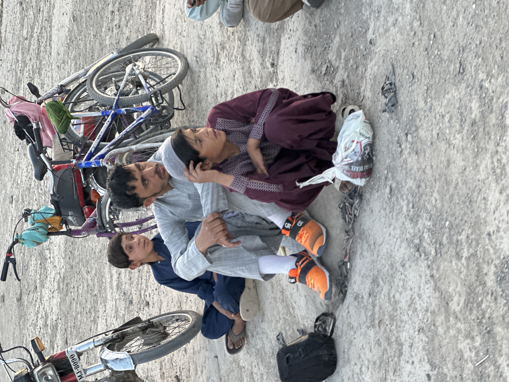 The image shows a group of people in an outdoor setting likely in or near Quetta City Tehsil, Pakistan, given the attire and general appearance. The ground is concrete or a similar hard, rough surface.
Three individuals are prominently featured: a man wearing a light gray outfit, a person in a maroon and gray patterned outfit, and a younger individual dressed in a blue outfit. The man in gray is sitting on the ground with the person in maroon leaning against him. The individual in blue is seated next to them.
Several bicycles are piled together nearby, and a motorcycle is also present. A shopping bag and a black bag are on the ground near the seated individuals. The overall scene suggests a moment of rest or gathering.