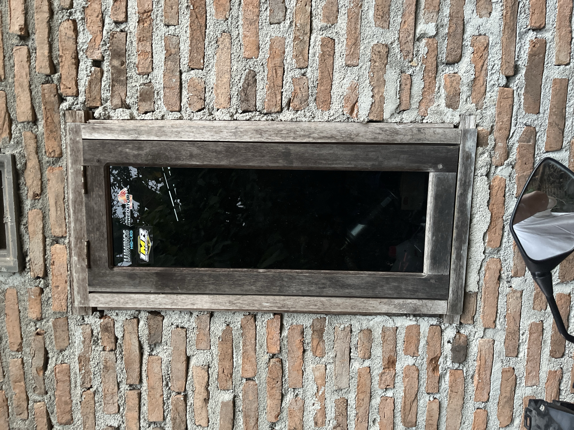 A vertical, medium shot captures a weathered wooden window set into a rough brick wall. The window frame is made of aged, graying wood, and the glass pane is dark and reflective, showing a blurred image of trees and a sticker that reads "LUMINOS HID-LED." Another sticker, partially obscured, appears to be a racing logo. Below the window, the handlebars and a side mirror of a motorcycle are visible, reflecting a white garment, possibly a shirt. The brick wall is constructed of reddish-brown bricks with rough gray mortar, characteristic of many buildings in Kebumen, Indonesia, suggesting a rural or semi-urban setting. The overall impression is one of a rustic, unfinished building, possibly a home or a small shop, in the Indonesian city of Kebumen.