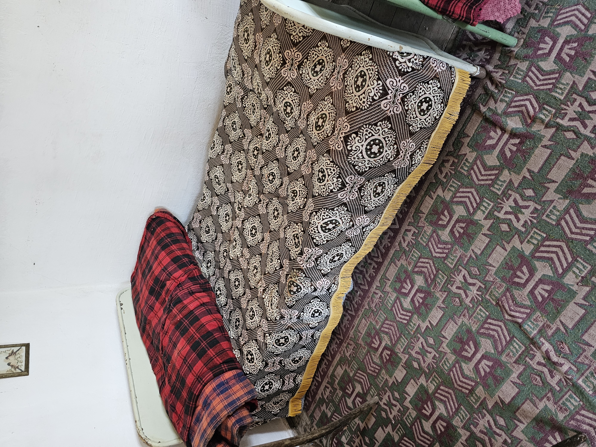 The image depicts a single bed situated in a room in Smolnik, Bulgaria. The bed features a light, possibly pale green or off-white, metal frame showing signs of age and wear on the head and foot boards. The mattress is covered by a patterned bedspread or skirt, which is dark brown or black with intricate cream-colored, curvilinear, and geometric motifs, interspersed with subtle reddish-pink accents. A yellow, fringed trim runs along the visible bottom edge of this bed covering.
On top of the bed, a large, rolled or folded blanket displays a prominent red and black buffalo plaid pattern. Beneath this, a section of another blanket or fabric with an orange and blue plaid design is visible. The walls of the room are painted a textured white, exhibiting some minor discoloration and marks. A small, framed picture, depicting two birds on a branch, hangs on the upper left wall.
The floor is covered by a woven rug or carpet featuring a geometric pattern composed of shades of green, muted purple, and light brown/beige. The pattern includes angular shapes resembling arrows, chevrons, and abstract symbols. Partially visible on the far left is a slender, dark wooden leg, likely belonging to another piece of furniture. On the far right, a portion of a furniture leg painted a similar pale green to the bed frame is visible, alongside a glimpse of red and pink plaid fabric. No individuals are present in the scene.
