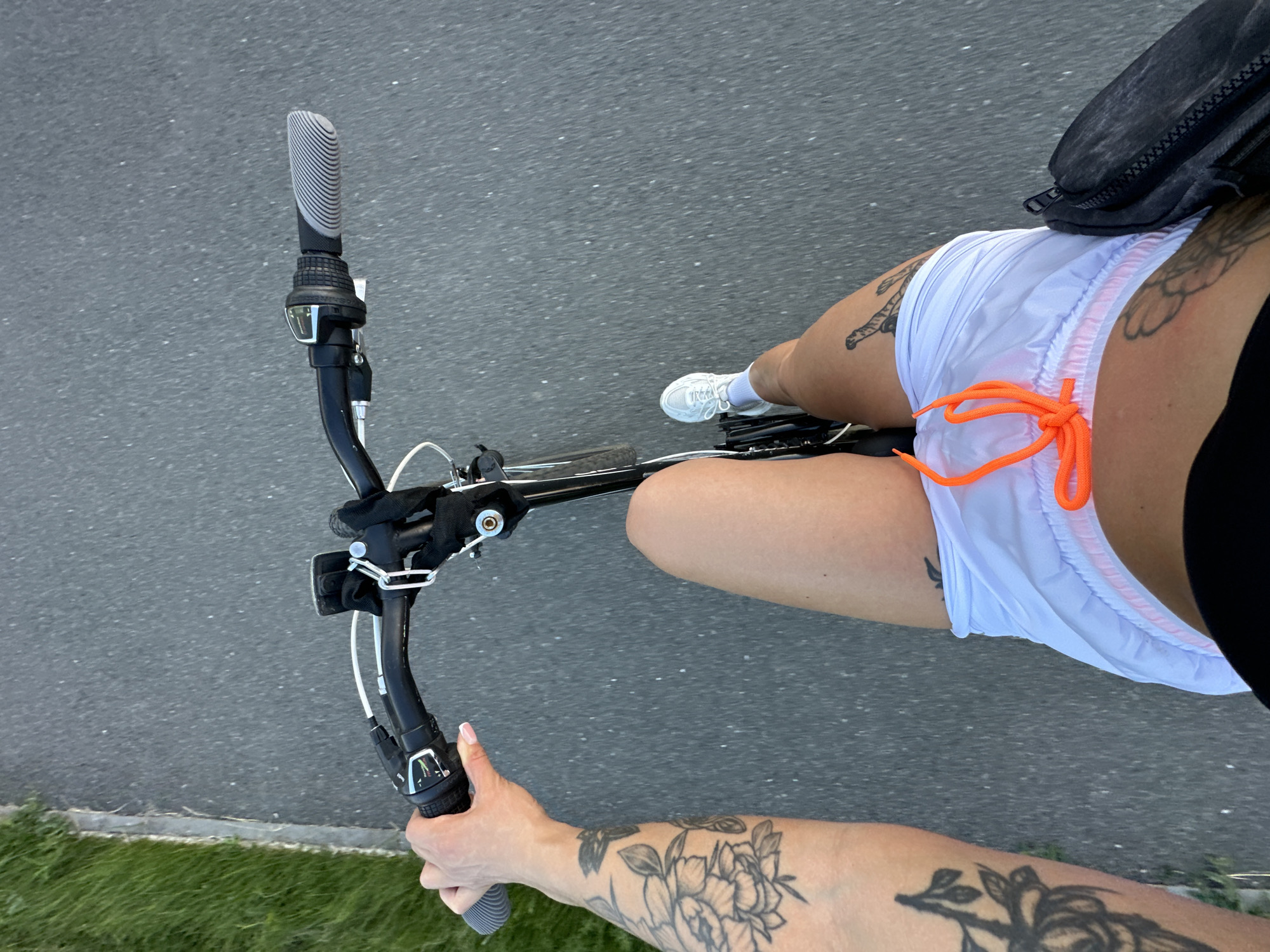 The image shows a first-person perspective of a person riding a bicycle on a paved path, likely in Yuzhnouralsk, Russia, given the context. The person is wearing white sneakers, white shorts with orange drawstrings, and a black top. They have extensive tattoos on their left arm and legs. The bicycle has black handlebars with grey grips and a gear shifter. To the left of the path is a strip of green grass. The path appears to be asphalt. A dark bag is attached to the bicycle, possibly a fanny pack or saddlebag. The image is taken from a high angle, looking down at the rider and the bike.