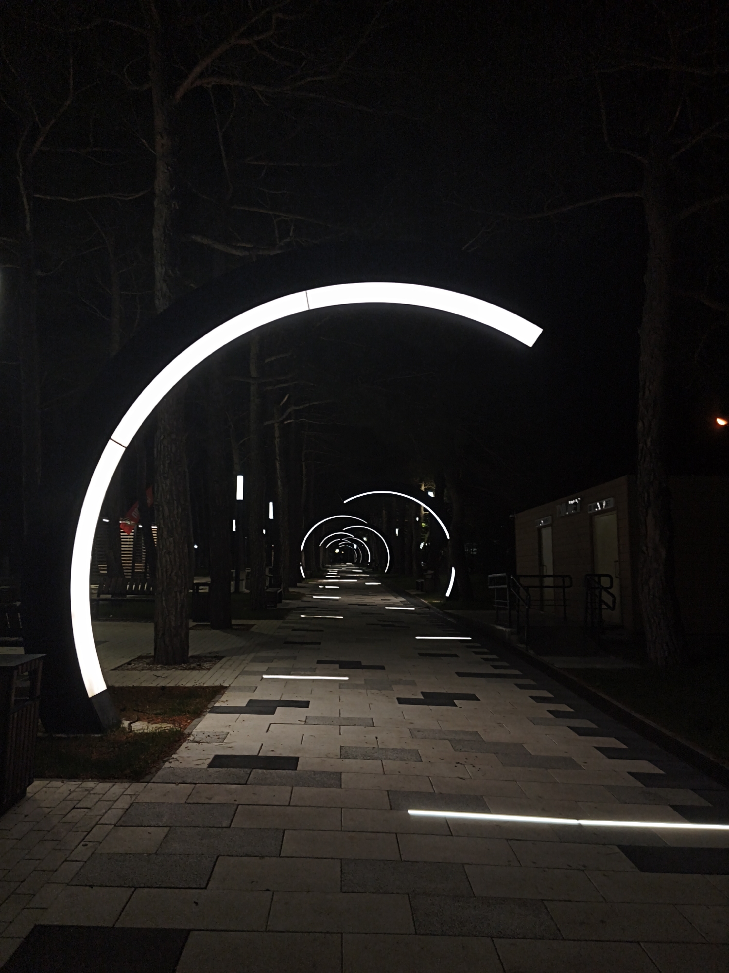 The image depicts an illuminated walkway at night in Myskhako, Russia. The walkway is paved with grey rectangular stones and features embedded horizontal light bars. Symmetrical arcs of white light are positioned along the path, creating a tunnel effect. Tall, dark trees flank the walkway. A small building is visible to the right of the walkway. Benches can be seen to the left. The image is dark, with the light features drawing attention to the perspective.