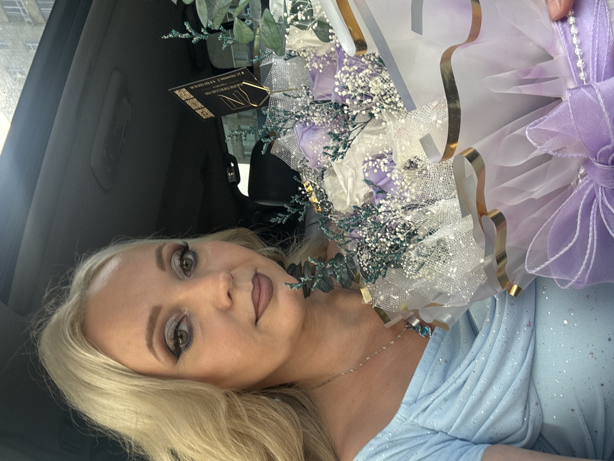 The image shows a blonde woman sitting in a car, holding a bouquet of purple and white roses. She is wearing a light blue, sparkling dress and a silver necklace with a blue pendant. The bouquet is wrapped in white and purple paper, with a lavender ribbon and pearl accents. A black card with gold lettering is attached to the bouquet, likely a gift card. The background is dark, suggesting the interior of a car, with a hint of the city skyline visible through the car windows. The woman is looking at the camera with a neutral expression. The scene is set in the city of Komsomolsk-on-Amur, Russia.