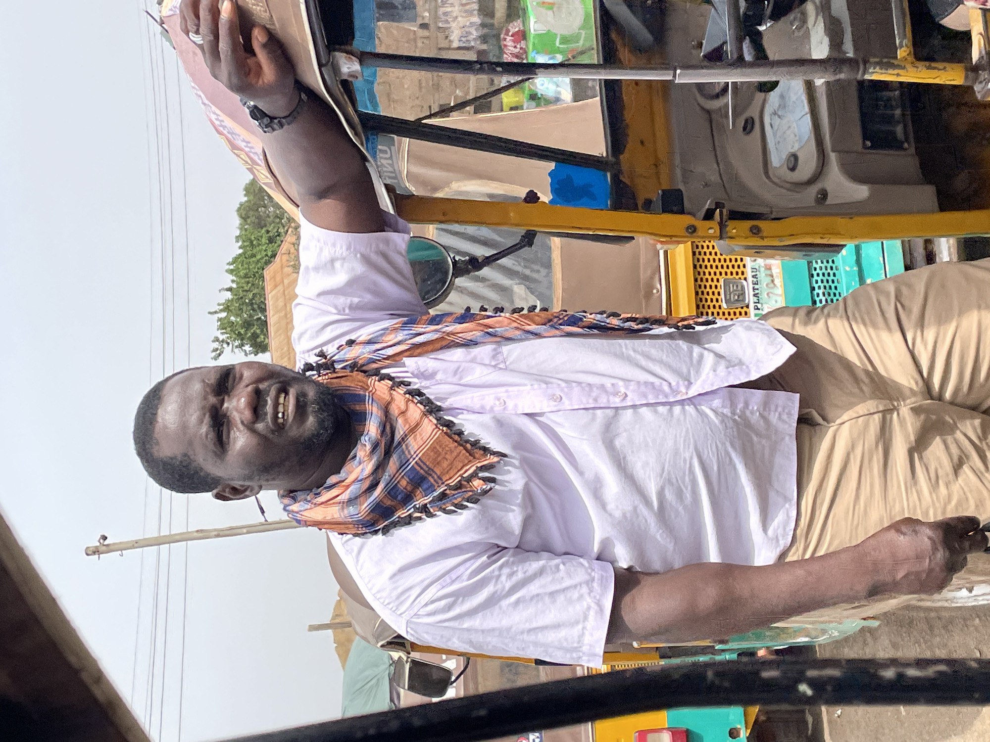 A man is the primary subject, appearing to be riding in or next to a yellow vehicle, possibly a taxi or similar transportation in the city of Jos, Nigeria. He is wearing a light-colored shirt with a checkered scarf draped around his neck, and khaki pants. The man is dark-skinned and has a black bracelet on his wrist and a ring on his finger. The vehicle is partially visible. Utility poles are in the background. The image is taken during the day with bright sunlight.
