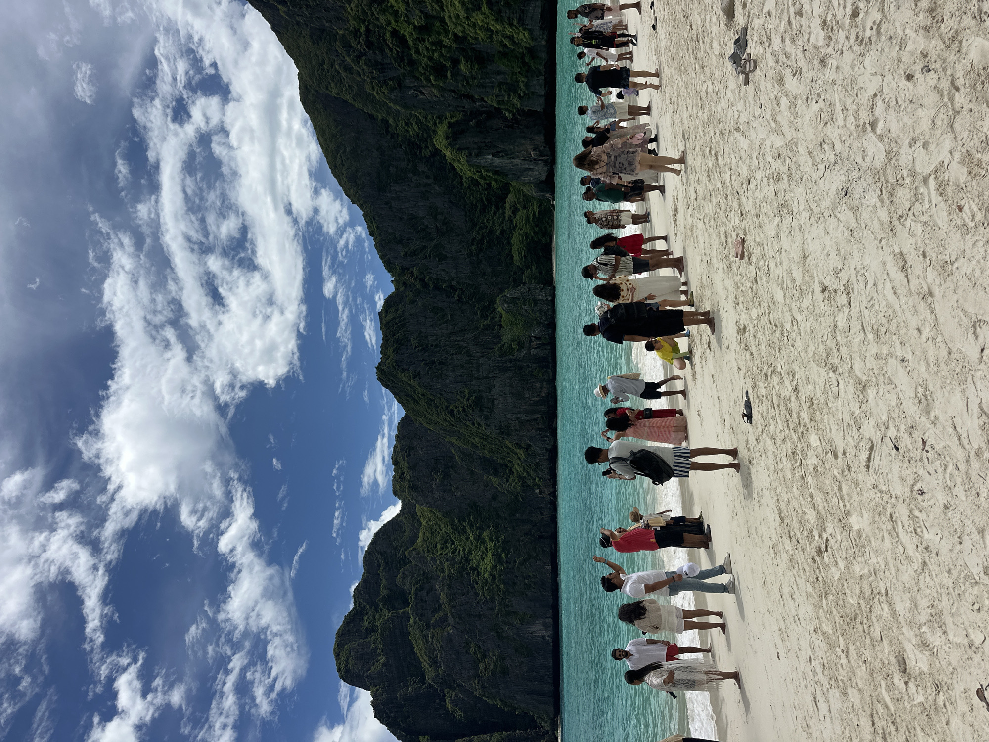 A crowd of people stand on a sandy beach in front of a body of turquoise water and a backdrop of large, vegetated cliffs. The sky above is blue with scattered white clouds. Many of the people are looking out towards the water or at the cliffs, while some are taking pictures. The scene appears to be a popular tourist destination, possibly Maya Bay in Krabi province, near Ao Nang, Thailand.