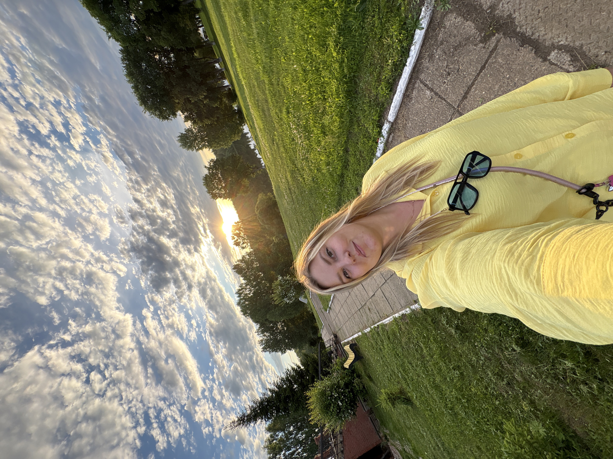 This is a selfie of a blonde woman wearing a bright yellow dress and sunglasses. She is outdoors, with a lush green lawn and trees visible in the background. The sun is setting, casting a warm glow on the scene and creating dramatic clouds in the sky. The woman's expression is neutral, and she appears relaxed. The location is Zavod Mikhailovskii, Russia. The time of day is sunset. The weather appears to be pleasant. There are no visible text elements in the image.