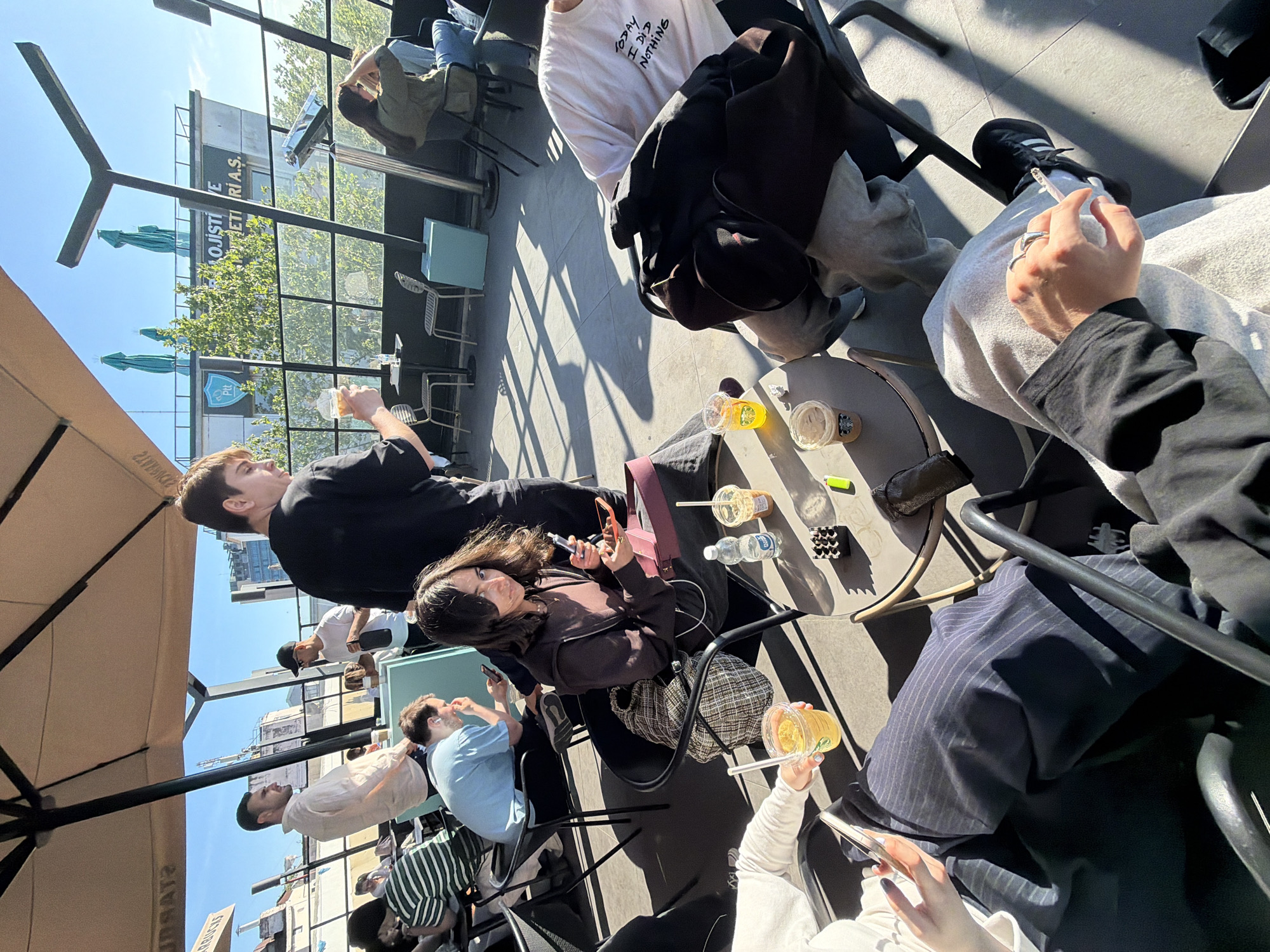 The image shows an outdoor scene at a Starbucks in Kadıköy, Turkey. Several people are seated at tables, some with drinks and personal items like phones, bags, and lighters on the tables. The architecture of the buildings across the street is visible through the outdoor patio's railing. The sky is blue.
