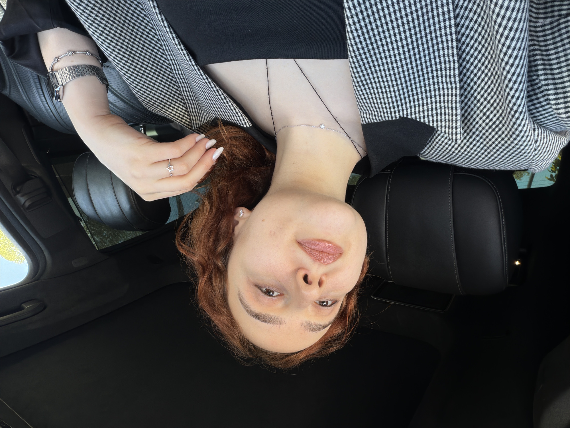 A young person with reddish-brown, wavy hair and fair skin is depicted, looking directly at the camera from an inverted perspective. She is reclining inside a car, with black leather seats and a dark headliner visible. She wears a black top under a black and white checkered blazer or jacket.
Notable accessories include a silver-colored watch with a metal band, a delicate silver bracelet with small stones or stars, and a silver ring with a star motif on her left hand. A thin silver necklace with a single clear stone pendant adorns her neck, and a small stud earring is visible in her right ear. Her fingernails are manicured with light polish and white tips.
The person has a neutral to slightly smiling expression, with light-colored eyes and makeup, including lip gloss. Bright daylight is visible through what appears to be a car window or sunroof. A smartphone is casually placed on the car seat beside her head. The image suggests a posed moment inside a vehicle, possibly taken from an overhead angle while the subject is lying down. There is no visible text in the image.