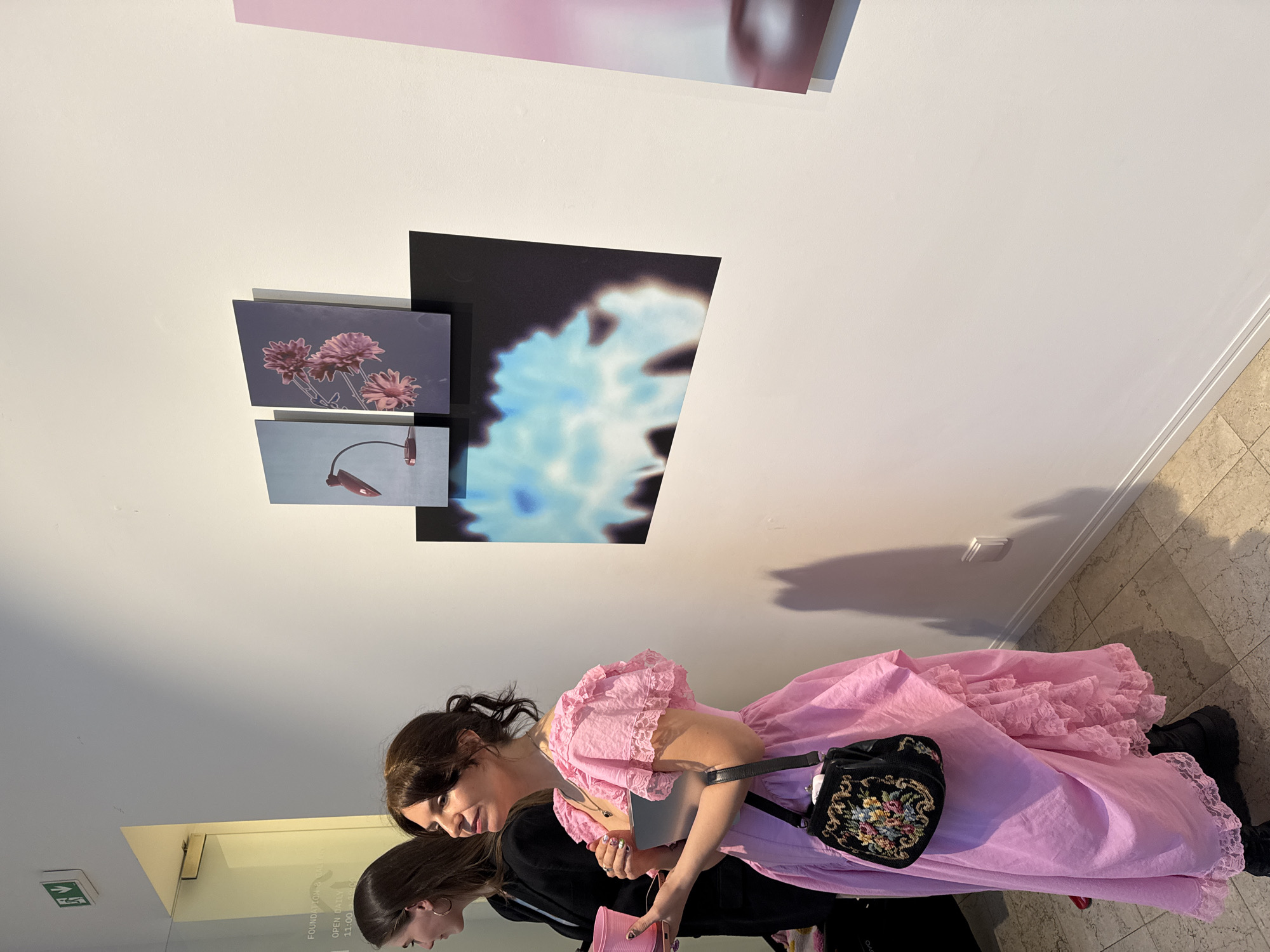 Two women stand in front of artwork displayed on a white wall in what appears to be an art gallery or museum. The artwork includes a photograph of pink flowers, an object suspended from a cord, and a partially out-of-focus image of blue and white shapes. The woman closer to the camera is wearing a pink ruffled dress and a floral embroidered black purse. The second woman is wearing a black jacket and holding a pink object. The floor appears to be tiled. The scene is in a well-lit indoor setting, presumably the city of Sofia, Bulgaria.