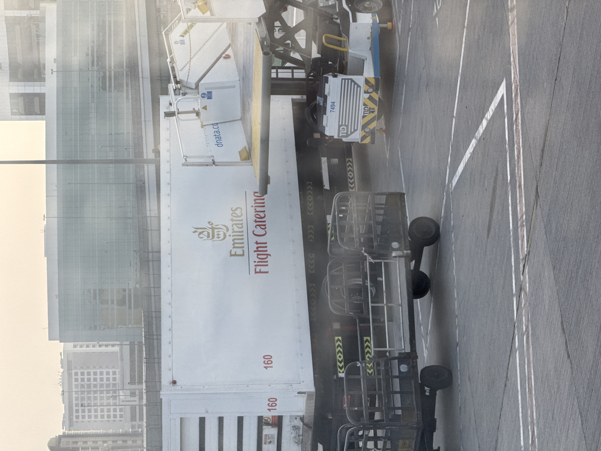 This image captures an airport apron scene in Dubai, United Arab Emirates, likely at Dubai International Airport (DXB). In the foreground, a catering truck with "Emirates Flight Catering" branding is parked, with two "160" numbers visible on its side. A luggage cart is positioned in front of the truck. To the right, a service vehicle with the number "7494" and the "TLD" logo is attached to a mobile stairs unit. The background shows airport terminal buildings, one with a distinctive modern glass facade, and other airport infrastructure. The overall lighting suggests it is daytime, possibly early morning or late afternoon, given the soft light and slight haze.