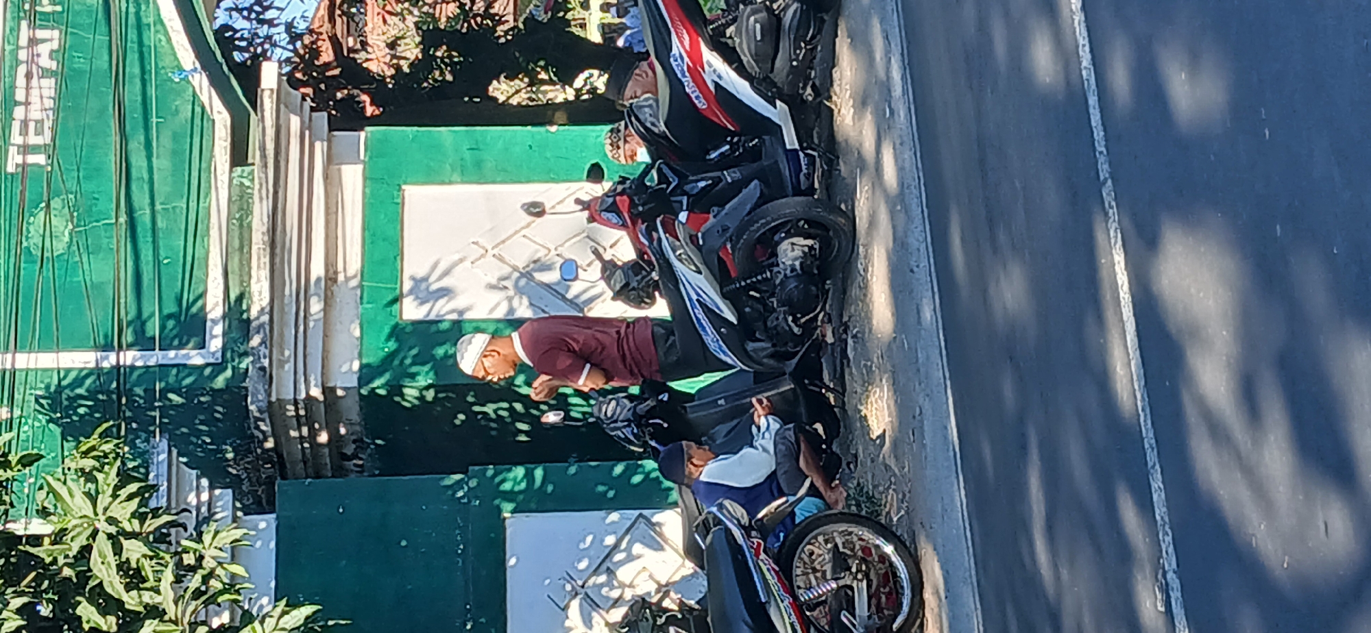 The image depicts a street scene in Kediri, Indonesia, featuring several motorcycles parked alongside a building with a green facade. The building has decorative white molding and a sign with text that appears to be "TEMPAT". A man wearing a maroon shirt and a white peci (cap) stands near the motorcycles, with his hands clasped in front of him, seemingly in prayer or contemplation. Another individual, a younger person wearing a blue and white jacket and a dark peci, is seated on the ground near a motorcycle, looking down at a phone. The motorcycles are parked on a dirt and gravel surface next to a paved road. The lighting suggests it is daytime, with shadows cast by trees. In the background, a few other people are visible, further down the street, along with more foliage.
