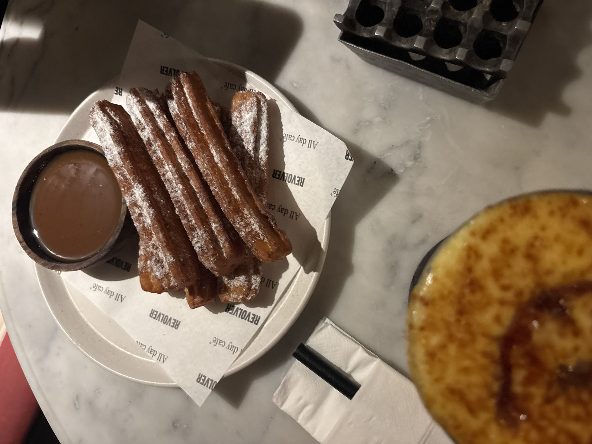 This overhead shot shows a plate of churros dusted with powdered sugar and a small bowl of dipping sauce, likely caramel, positioned on a white marble table. The churros are arranged artfully on a piece of paper with the text "REVOLVER All day cafe" printed repeatedly. To the right of the churros, a blurred close-up of a crème brûlée dessert is visible, with a dark syrup drizzled over its caramelized top. A dark straw rests on a white napkin next to the dessert. In the background, a metal object with multiple circular compartments, possibly an ashtray, is situated on the table. The lighting suggests it might be indoors, with shadows casting onto the marble surface, indicating either artificial light or a time of day with strong directional sunlight. The overall scene evokes a relaxed cafe setting, likely in North Kuta, Indonesia, where one might enjoy sweet treats and beverages.