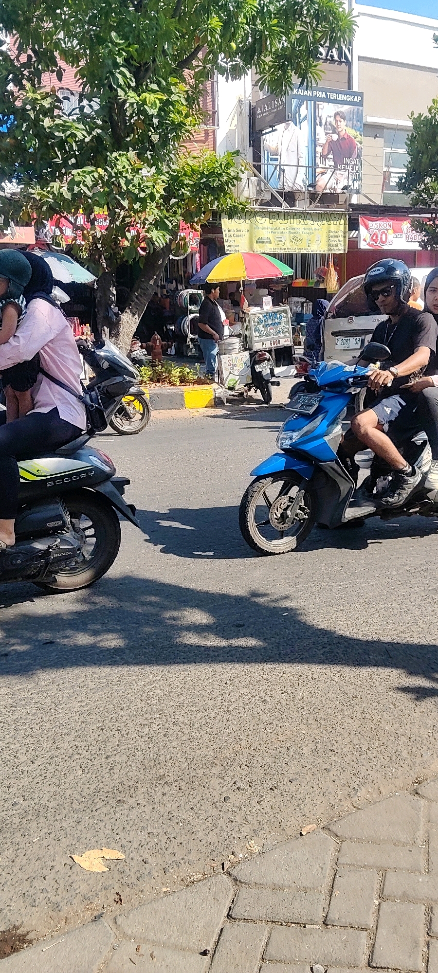 The image depicts a street scene in Cibatu, Indonesia, on a sunny day. In the foreground, two motorbikes are prominent. A black scooter, labeled "SCOOPY," is partially visible on the left, with its rear wheel and a portion of its body showing. The rider's leg, wearing black pants and a white sandal, is also visible. On the right, a bright blue motorbike is more fully shown. It is ridden by a man wearing a black helmet, sunglasses, a black t-shirt, and shorts. He is looking to his right, and a woman with long dark hair, wearing a black top, is seated behind him.
The background reveals a bustling street environment. Several buildings line the street, one featuring a large advertisement with a man in a suit and text in Indonesian. Other shops or stalls are visible, with various goods displayed, including what appear to be kitchen appliances and cookware. A colorful umbrella shades a small cart, where a man in a black shirt and jeans stands. In front of the cart is a large cylindrical container. Another motorbike, a yellow and black model, is parked on the side. The road surface is asphalt, with some cobblestone paving visible at the edge of the sidewalk. Trees with green foliage provide some shade, and shadows are cast across the road. A fire hydrant is visible on the sidewalk.