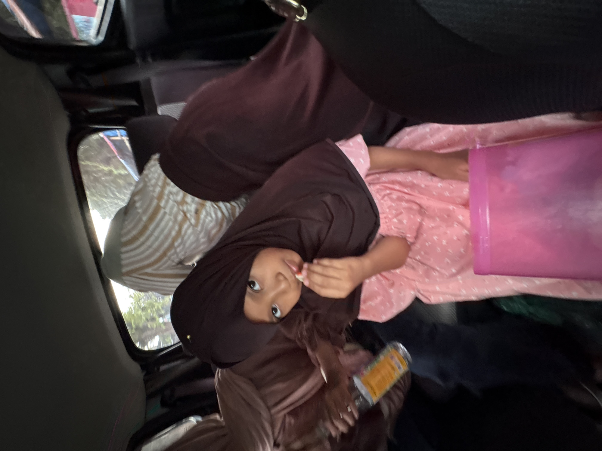 A young girl wearing a dark brown hijab and a pink dress with white polka dots is seated inside a vehicle. She is holding a small piece of food to her mouth. Behind her, another person, possibly an adult, is visible, also wearing a hijab. The interior of the vehicle appears to be a car, with seats and windows. The lighting suggests it is daytime, with trees and foliage visible outside the windows, indicating an outdoor environment, possibly along a road in Sumbersari, Indonesia. A pink container is placed in front of the girl.