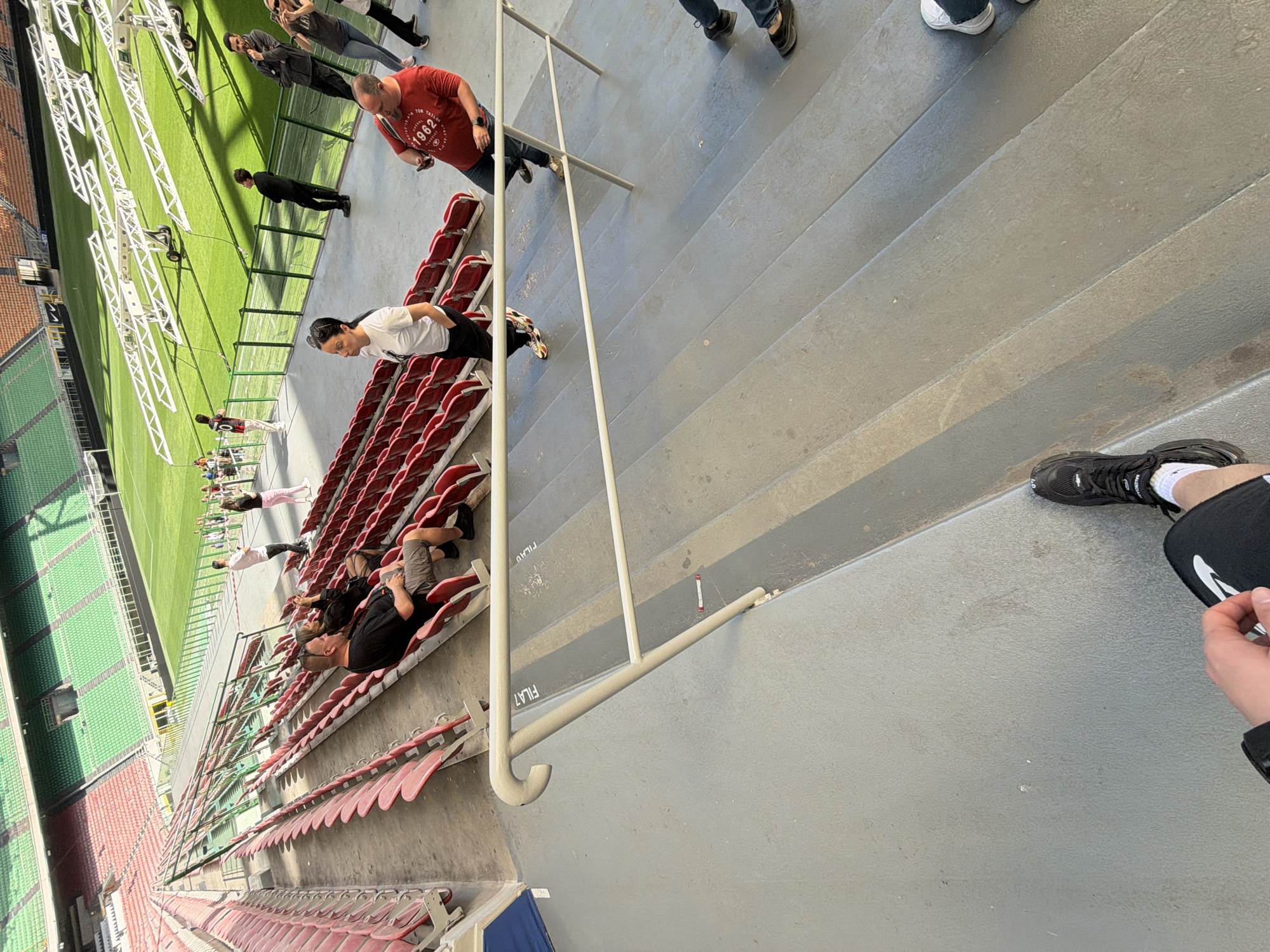The image depicts the interior of San Siro stadium in Milan, Italy. The view is from an elevated position on concrete steps, looking down towards the stadium. Several people are visible in the stands, some seated in red stadium chairs, others standing or walking. A metal railing runs parallel to the steps, separating the seating area from the walkway. The pitch is green, with white equipment structures along its edge. Some individuals are on the pitch. The seating arrangement extends downwards, suggesting a large stadium capacity.