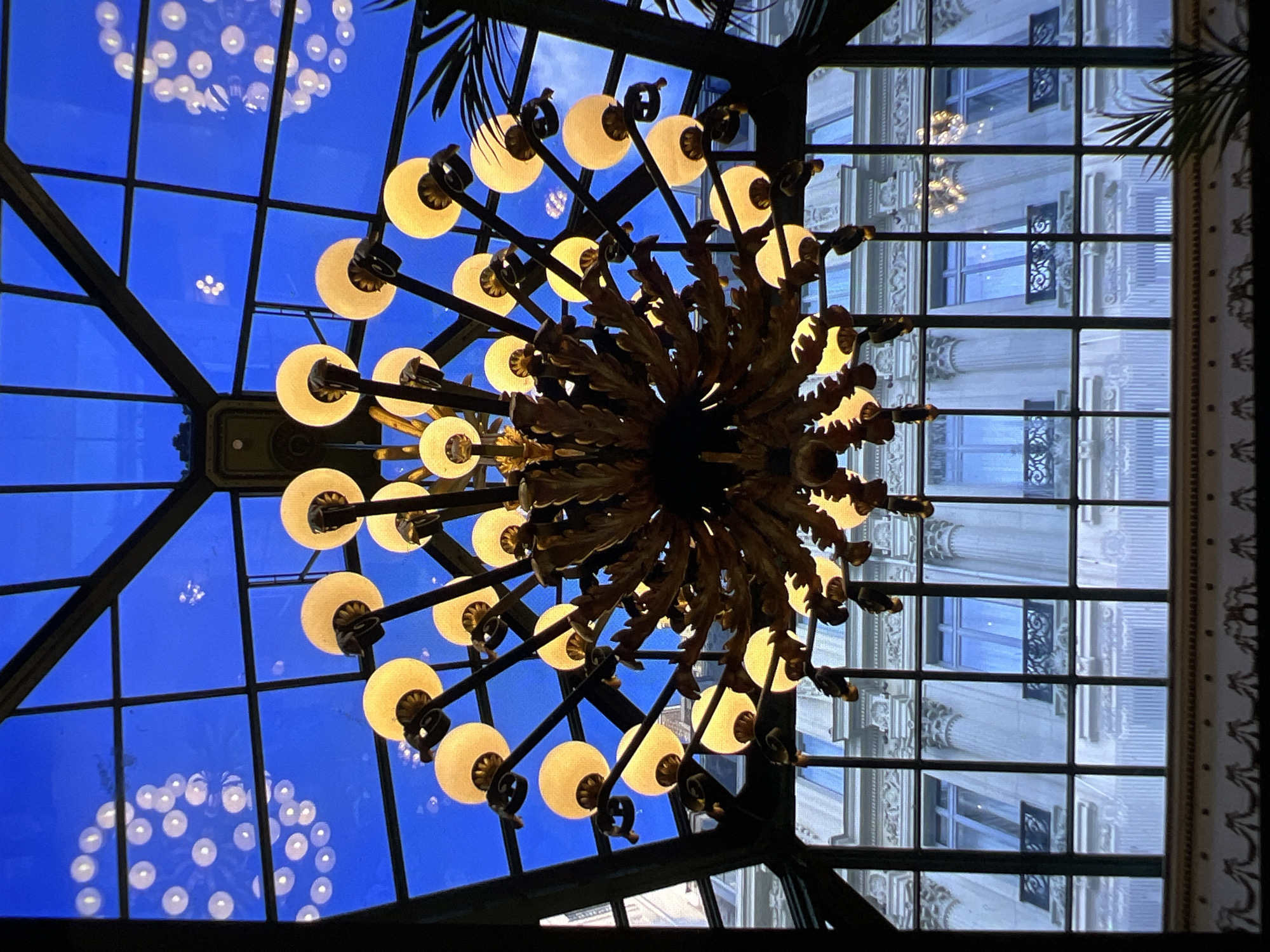 A chandelier with many lights hangs in front of a glass ceiling, which reflects more lights. Below the glass ceiling, there is a building with many windows and columns. The building's architectural style is neoclassical. The photo was taken in Verona, Italy.
