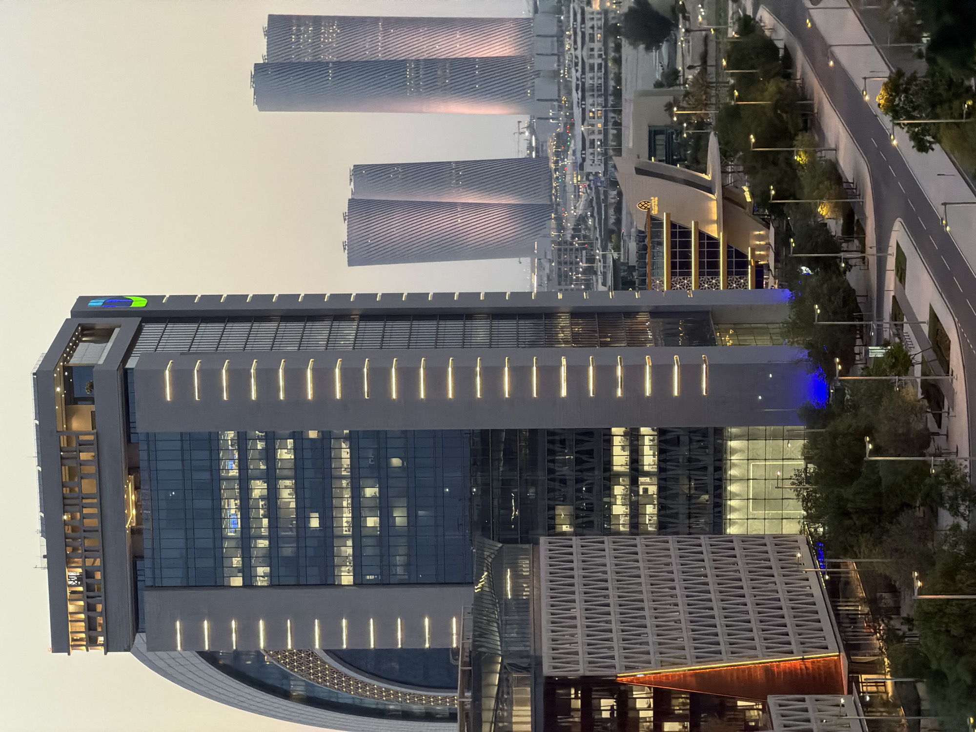 This is a vertically oriented, eye-level medium shot of a modern cityscape at dusk. The scene features several tall, sleek skyscrapers with distinctive architectural designs. The foreground is dominated by a large, multi-story building with a glass facade and illuminated interior sections. The building's exterior is accented with vertical light fixtures. To the left of this building, another structure with a curved, semi-circular base is visible. In the background, three pairs of identical, twisted skyscrapers rise towards the sky. These structures have a metallic sheen and are subtly illuminated by the fading sunlight, casting pinkish hues on their surfaces.
The surrounding environment includes a well-maintained road with streetlights, a sidewalk, and lush green trees, suggesting an urban park or landscaped area. The sky is a pale, hazy gradient of light orange and beige, indicating the transition from day to night. Lights are visible in the windows of the buildings, suggesting that people are inside working or present. The overall atmosphere is one of modern urban development and evening activity.
There is a visible logo on the top section of the foreground building that appears to be a stylized graphic with blue, green, and yellow colors. Text can be seen on a building in the mid-ground, though it is too small to read clearly. The time of day is dusk. The weather appears clear. The location is likely a modern, affluent urban center.
