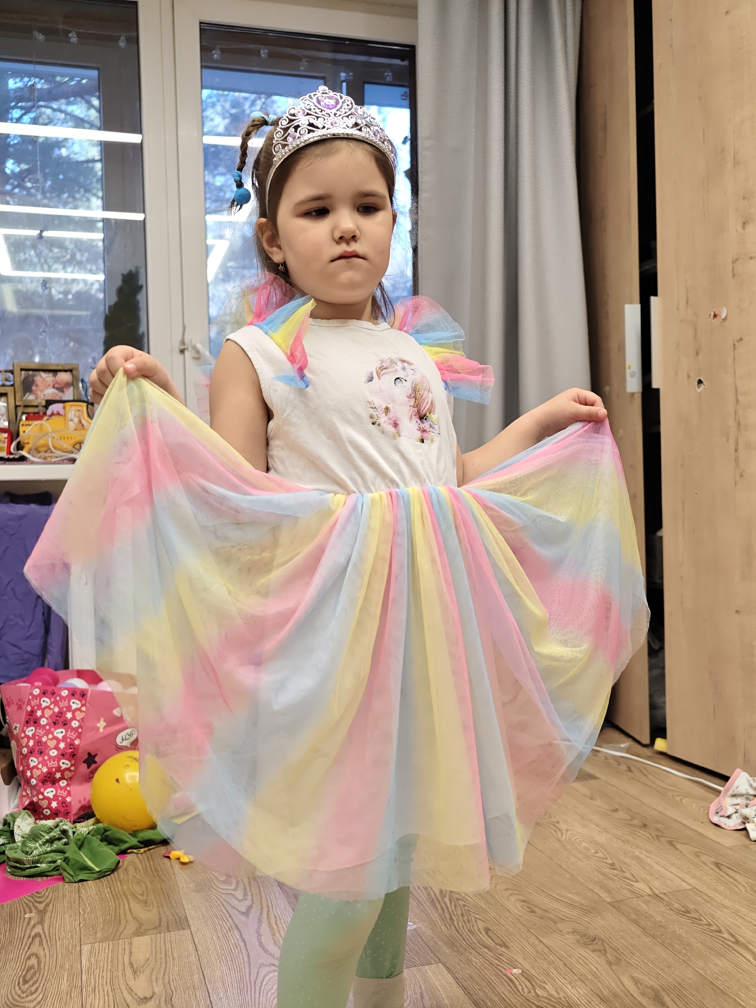 The image features a young girl, appearing to be between four and seven years old, dressed in a colorful costume indoors. She wears a sparkly silver tiara with purple jewels, a white short-sleeved top with a cartoon unicorn graphic on the chest, and a voluminous, layered tulle skirt in soft rainbow hues of pink, yellow, blue, and green. Matching colorful tulle ruffles are visible on her shoulders. She also has light green patterned leggings and her dark hair is partially braided with blue ties.
The girl stands centrally, holding out the sides of her skirt as if displaying it. Her expression is unsmiling and appears somewhat serious or contemplative.
The setting is a room with light wooden floorboards. To the left, a large window with horizontal blinds shows bare tree branches outside, suggesting a cooler season. Below the window, a shelf holds various items, including framed photographs and a yellow toy vehicle. A grey curtain is drawn over part of the window. On the floor in the foreground, there is a pink patterned bag, a yellow ball, and some green fabric. To the right, a light brown wooden wardrobe or cabinet is partially visible. The scene is illuminated by natural daylight coming from the window. No discernible text is visible in the image. The location context is Chaikovsky, Russia.