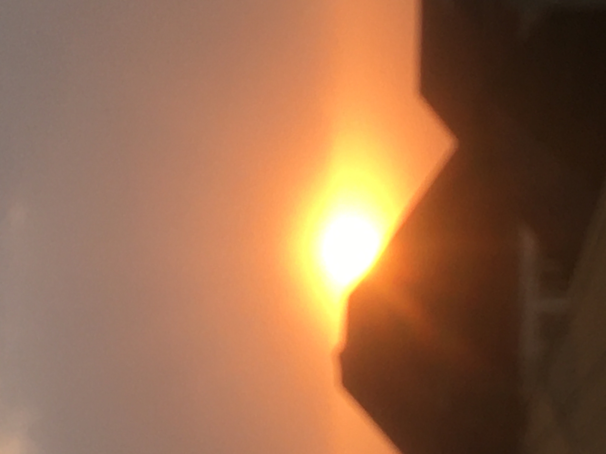 The image shows a blurred view of a sunrise or sunset in Dutsen Alhaji, Nigeria. A bright, glowing orange and yellow sun is partially obscured by a dark, indistinct building silhouette to the right. The sky is a hazy gradient of orange and gray.