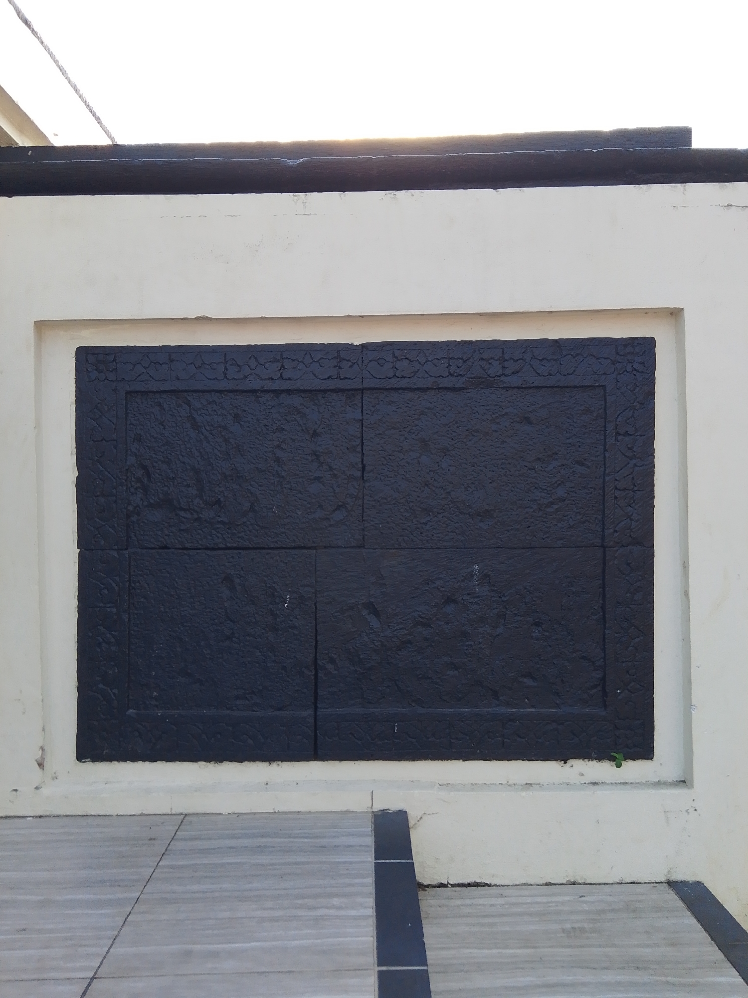 A wall of a building with a light cream color is visible. A rectangular dark-colored relief is embedded in the wall. The relief is divided into four square sections, with intricate patterns carved around its border and within the sections. The relief is framed by a lighter cream border that is flush with the wall. A dark-colored overhang runs along the top of the wall. In the foreground, a tiled pathway with gray and white tiles is visible. The scene appears to be outdoors, possibly in a public space or courtyard. The sky is bright and appears to be daytime. The location is identified as Demak, Indonesia.