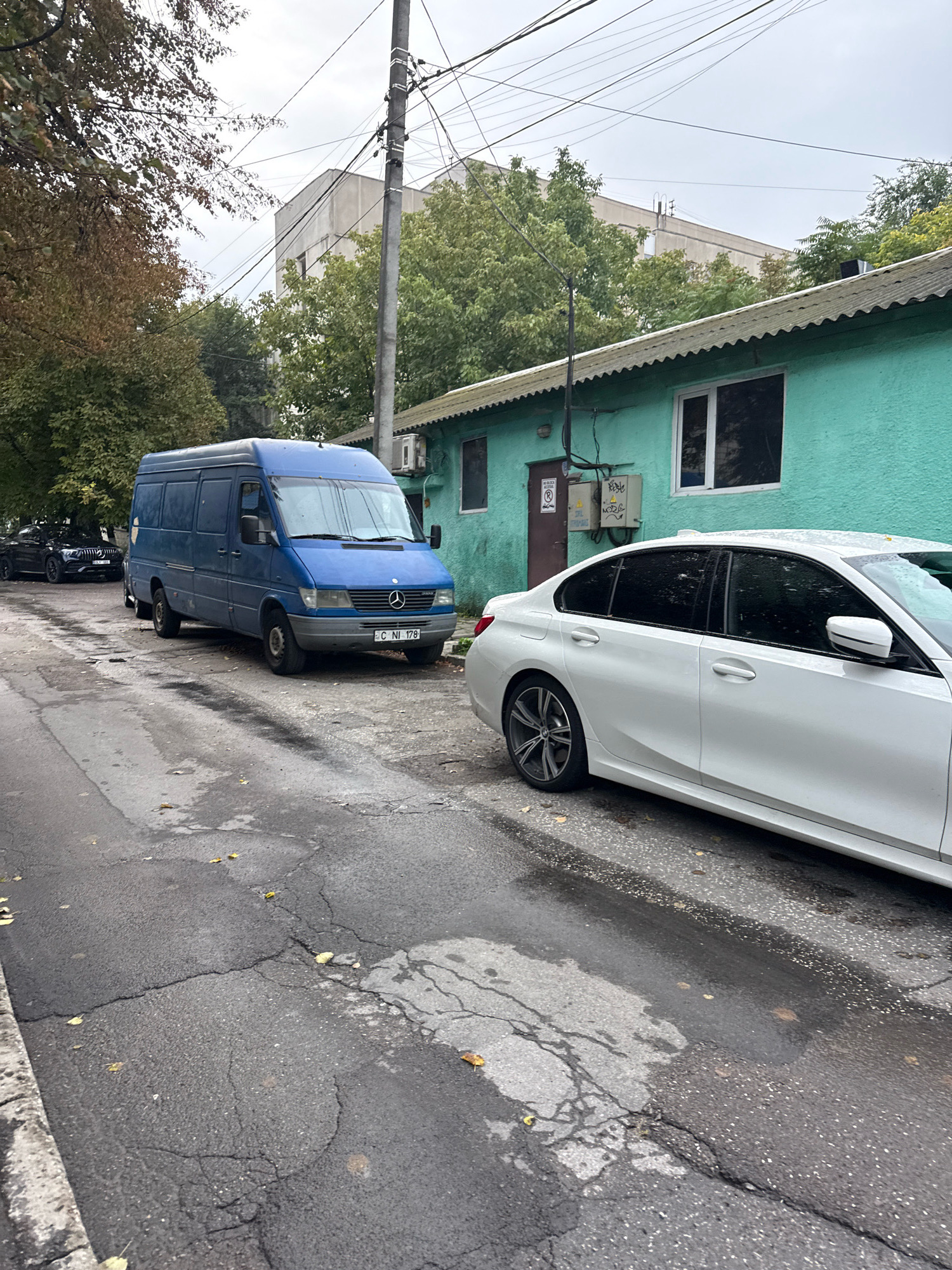 The image captures a street scene in Chișinău, Moldova, with a focus on parked vehicles and a building. In the foreground, a cracked asphalt road shows signs of wear and some puddles, with fallen leaves scattered around, suggesting an autumn or damp weather condition. To the right, the rear of a white sedan is visible, parked on the side of the road.
Dominating the midground is a blue Mercedes-Benz Sprinter van, parked on the left side of the street. The van's license plate is visible, reading "C-NI 178". Beside the van, further down the street, a black luxury sedan is also parked.
Behind the vehicles, a low, turquoise-colored building with a corrugated metal roof is prominent. It features windows and an air conditioning unit mounted on the exterior wall. A utility pole with numerous wires stretches across the scene above the building and trees. In the background, a multi-story concrete building can be seen, partially obscured by trees. The sky is overcast. The overall impression is that of a quiet urban residential or commercial street.