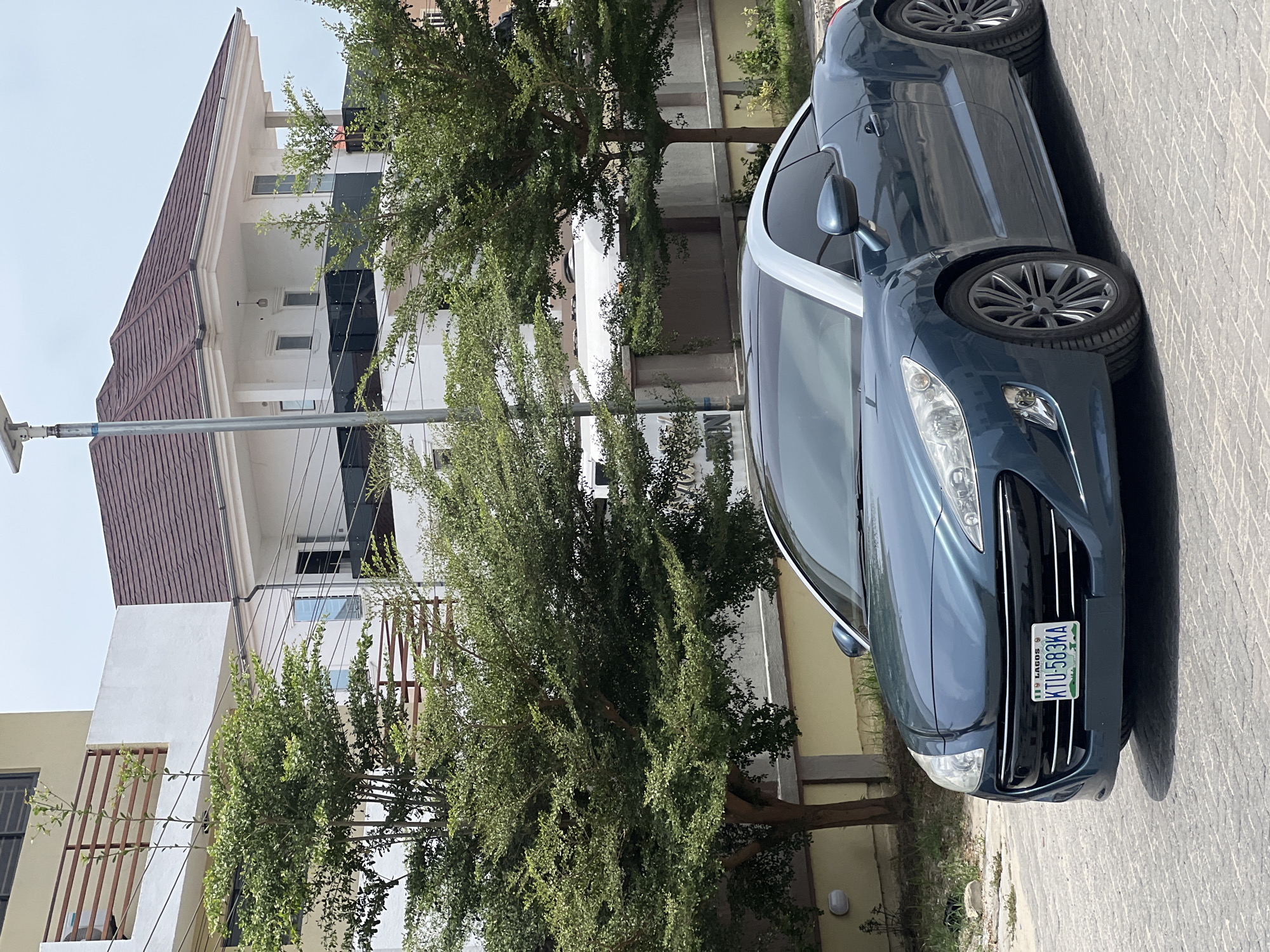The image captures an outdoor scene in Igboefon, Nigeria, featuring a dark blue car parked alongside a modern building and several trees under bright daylight. The car, visible from the front and side, appears to be a coupé or a sleek sedan, possibly a Peugeot, with intricate wheel designs and clear headlights. Its front license plate is clearly visible, displaying "LAGOS" at the top and "KTU-583KA" below it, alongside a small Nigerian flag symbol. The car is parked on a light-colored paved surface, casting prominent shadows that indicate a sunny day.
Behind the car, a contemporary multi-story building dominates the background. It is predominantly white with sections of dark paneling or tinted windows and features a brown or dark-tiled gabled roof. Balconies with dark railings are visible on the lower floors. Several lush green trees are scattered throughout the scene, partially obscuring the building and adding a natural element to the urban environment. A streetlight pole can be seen on the far left. The scene depicts a quiet moment in what appears to be a developed residential or commercial area. No people or specific activities are visible, emphasizing the stillness of the street.