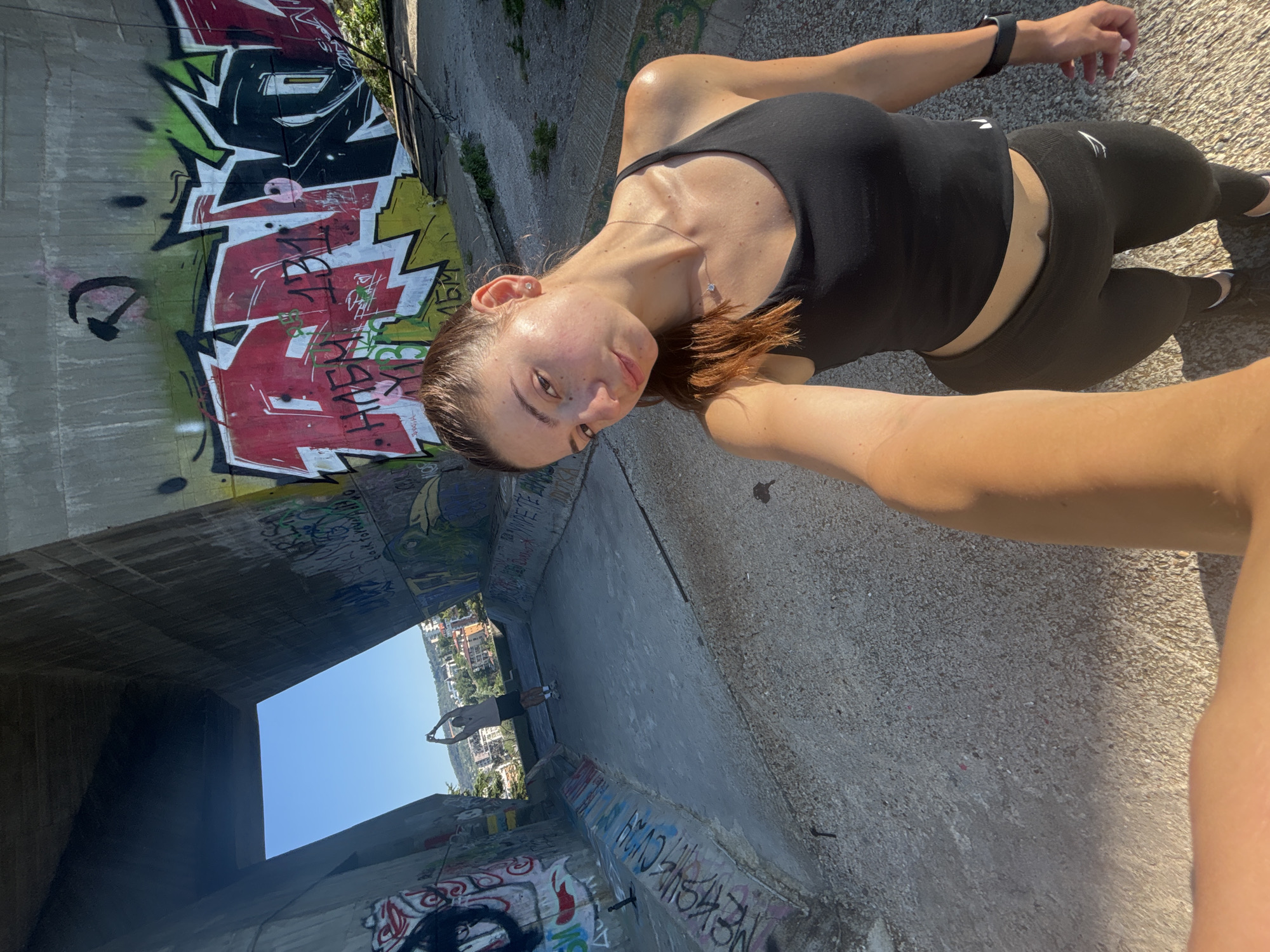 The image captures a sunny day under a concrete overpass in Varna, Bulgaria, adorned with graffiti. A young woman in a black tank top and leggings is in the foreground, taking a selfie. Her hair is pulled back in a ponytail, and she wears a necklace and earrings. Behind her, a person in athletic wear stands with arms raised, seemingly stretching or exercising. The overpass walls are covered in colorful graffiti, and the ground is concrete. The background shows a cityscape with buildings and trees under a clear blue sky.
